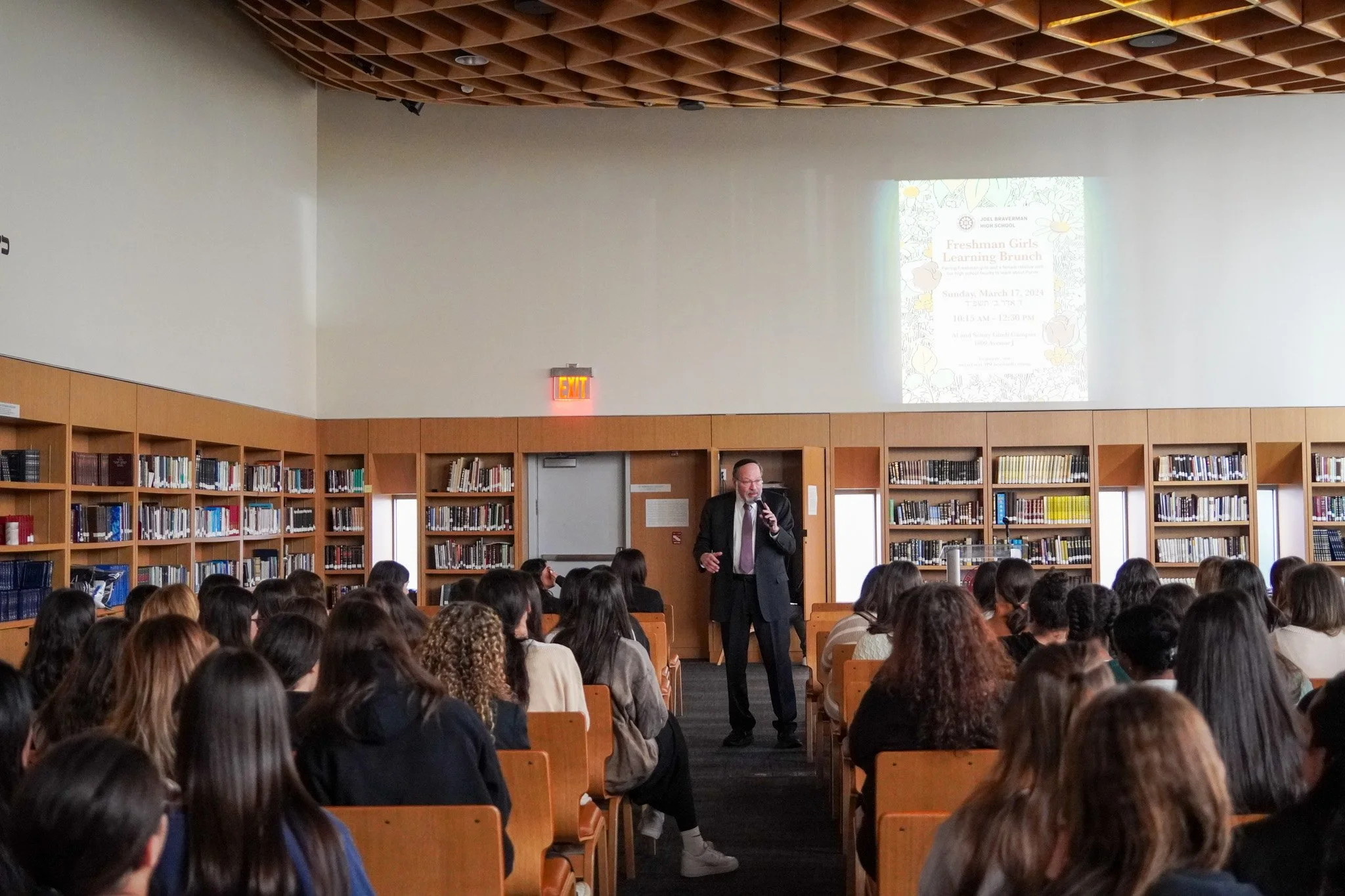 A speaker in a black suit and tie stands in front of an audience of high school students in a library, giving a presentation for a Freshman Girls Learning Brunch event.