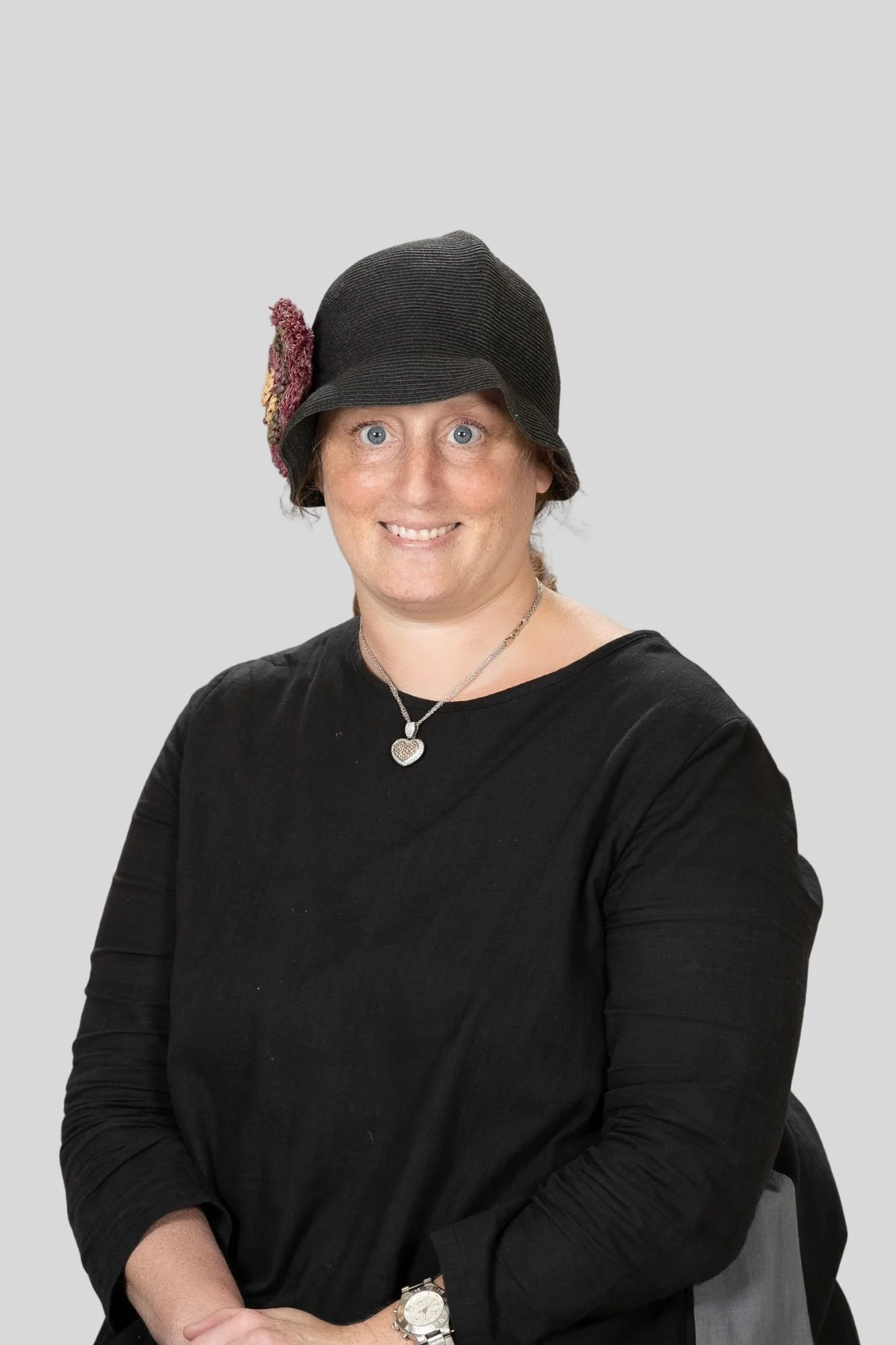 A woman with blue eyes, wearing a black long-sleeve shirt, a necklace with a heart-shaped pendant, a silver watch, and a black hat with a pink decorative flower, smiling at the camera.