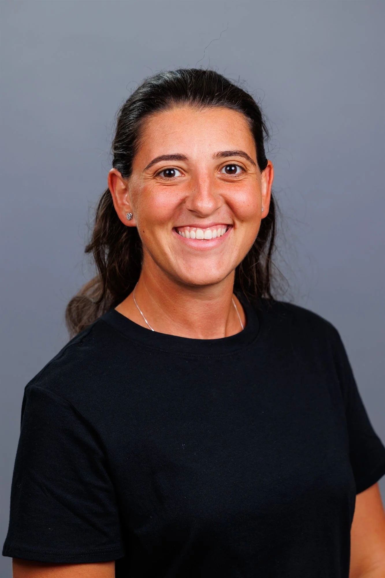 Headshot of a woman with dark hair pulled back, wearing a black shirt, smiling against a plain gray background.