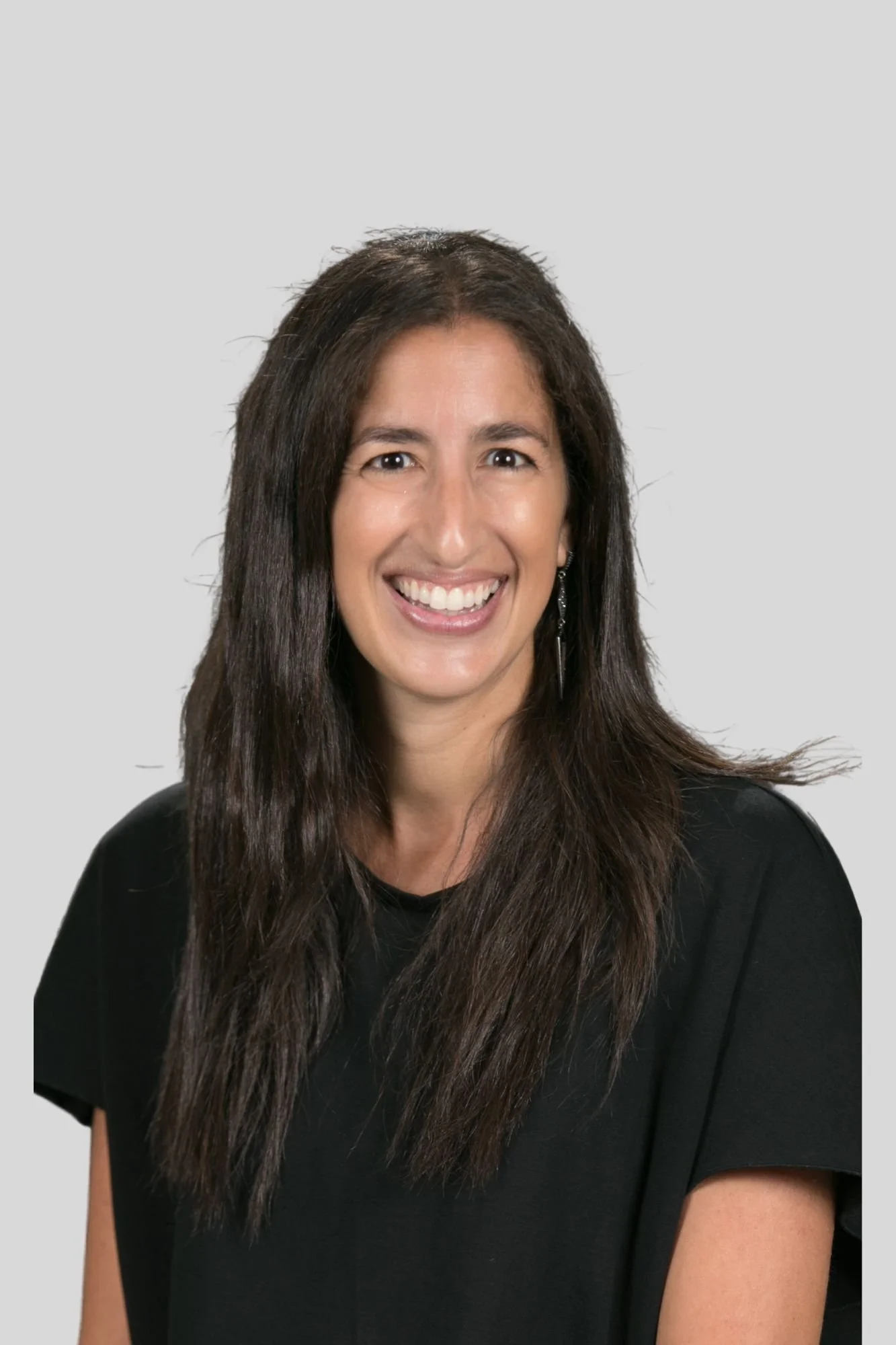 A woman with long dark hair wearing a black shirt, smiling against a light gray background.