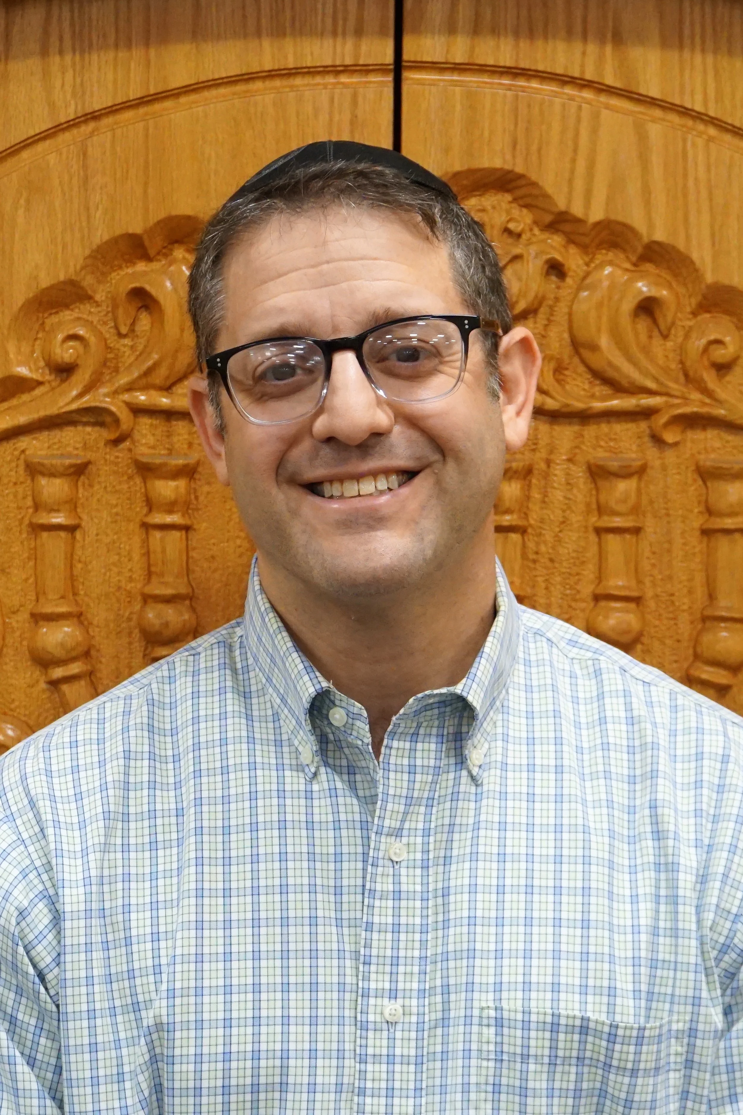 Smiling man wearing glasses and a kippah in front of a carved wooden headboard.