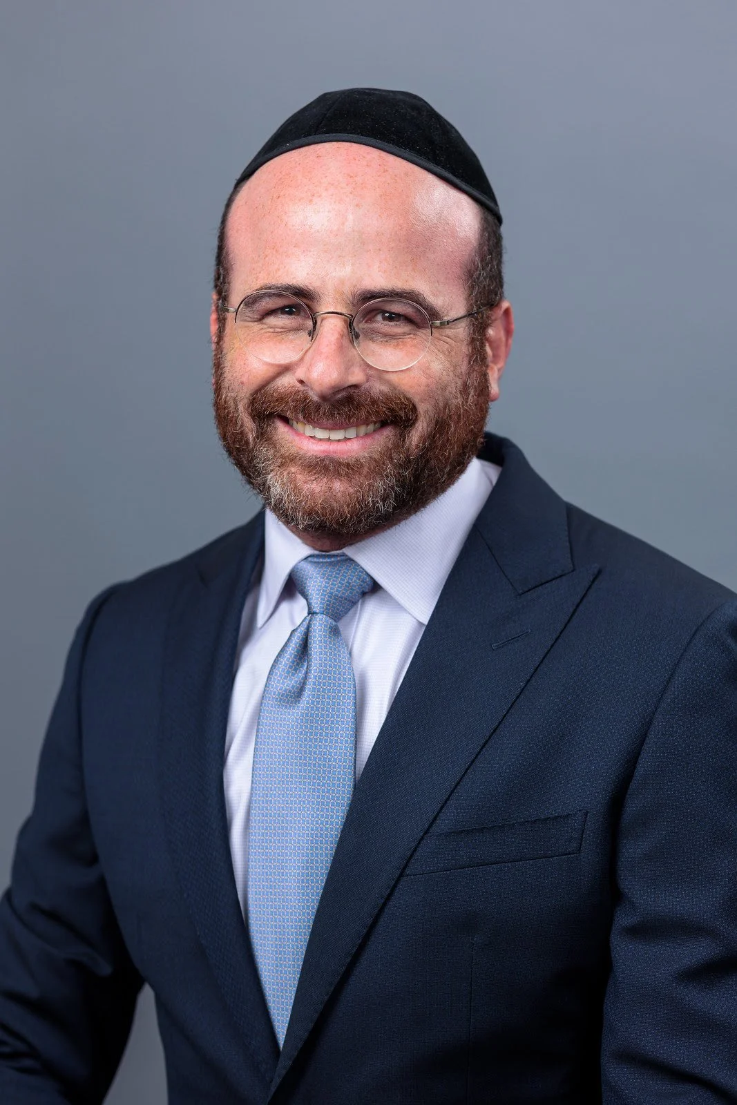 Portrait of a man with a beard, glasses, and a kippah, wearing a navy suit and light blue tie, smiling against a gray background.