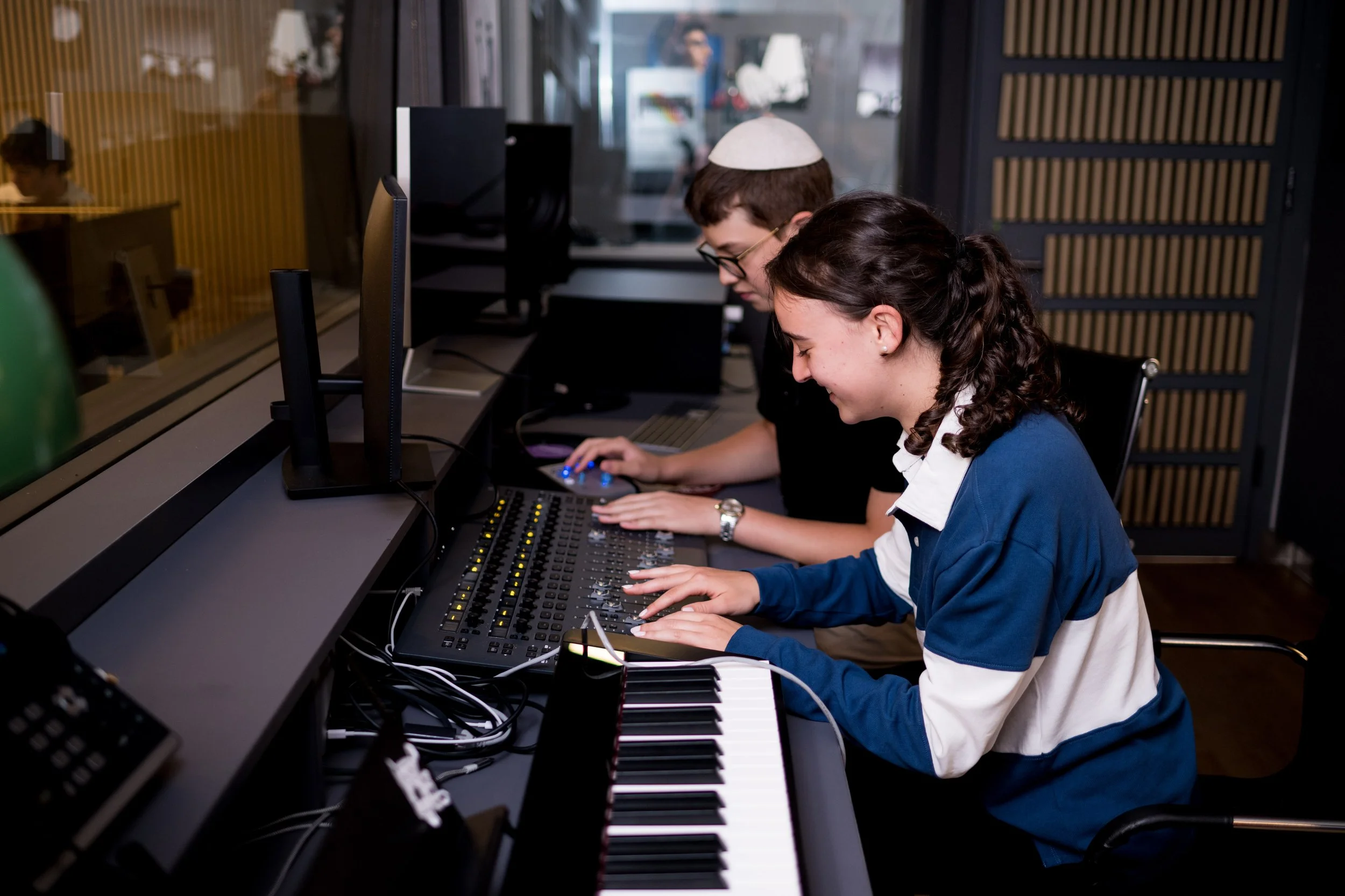 Two young people are working on music production equipment in a recording studio, with a keyboard and mixing console.