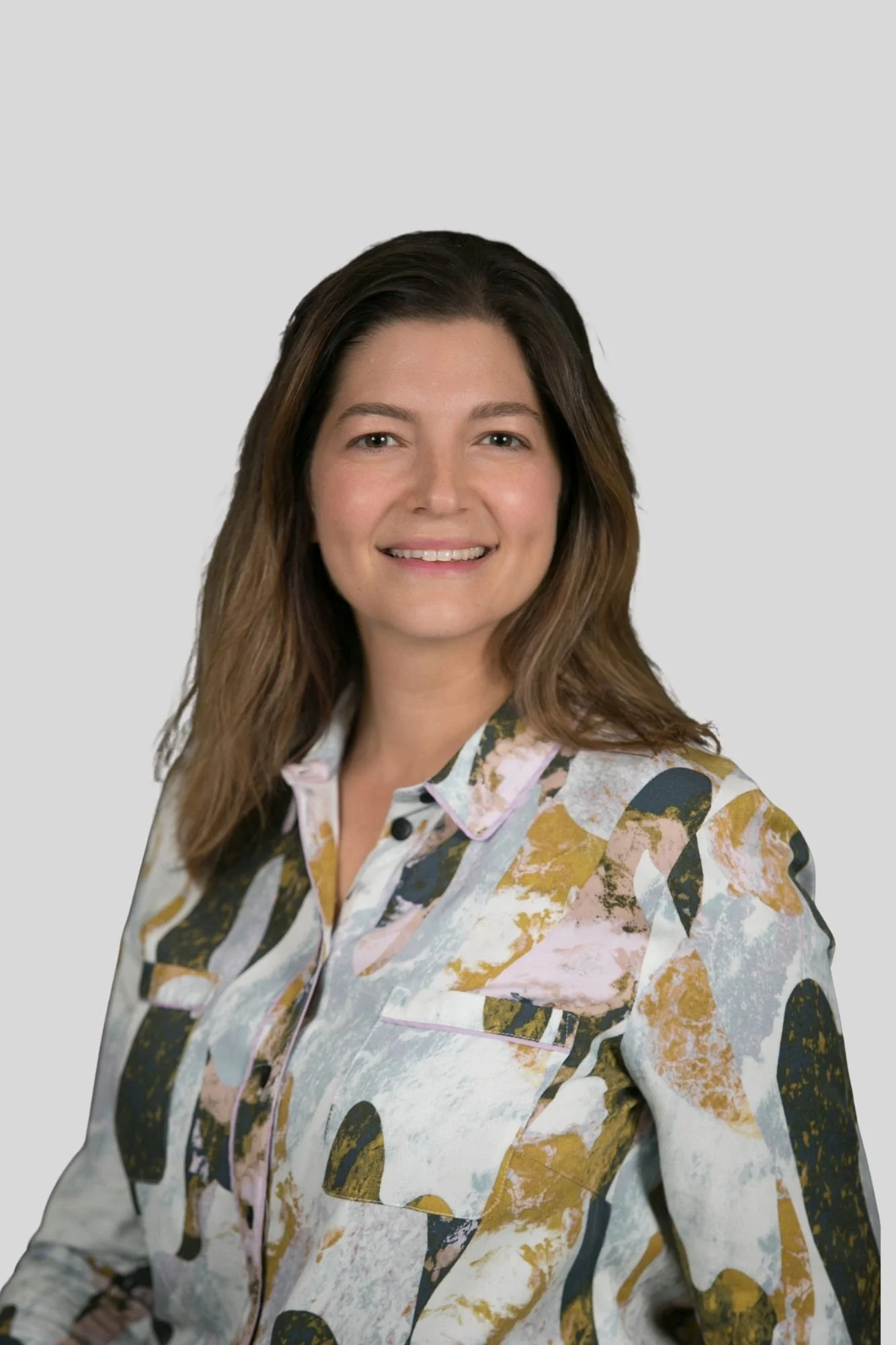 Portrait of a woman with long, wavy brown hair smiling, wearing a colorful, abstract-patterned button-up shirt, against a plain light gray background.
