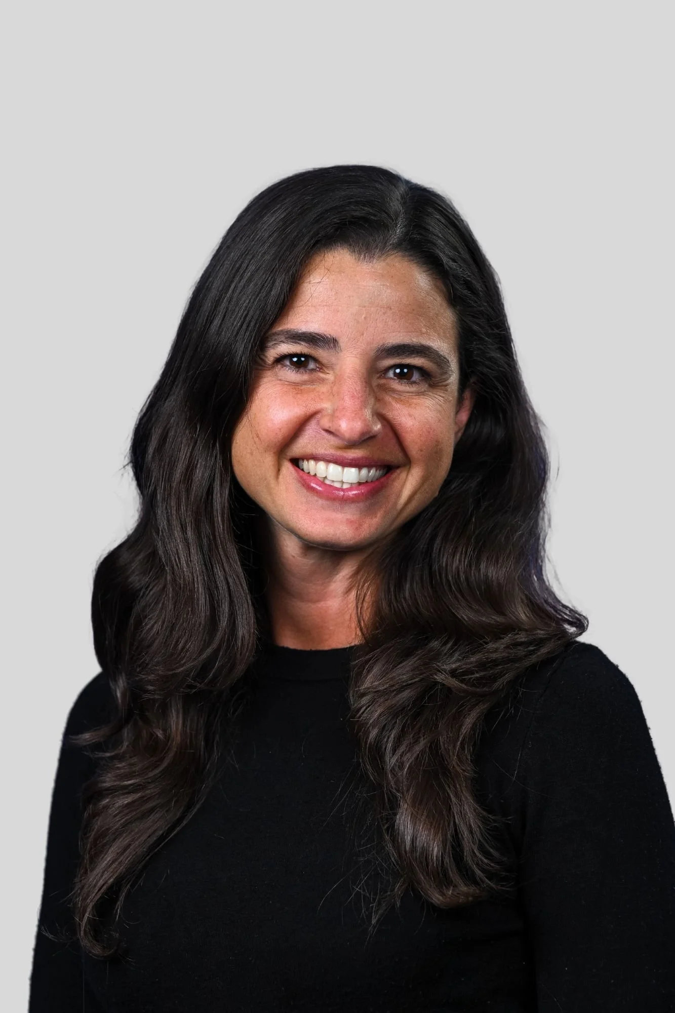 Close-up portrait of a smiling woman with long, wavy dark hair, wearing a black top, against a plain gray background.