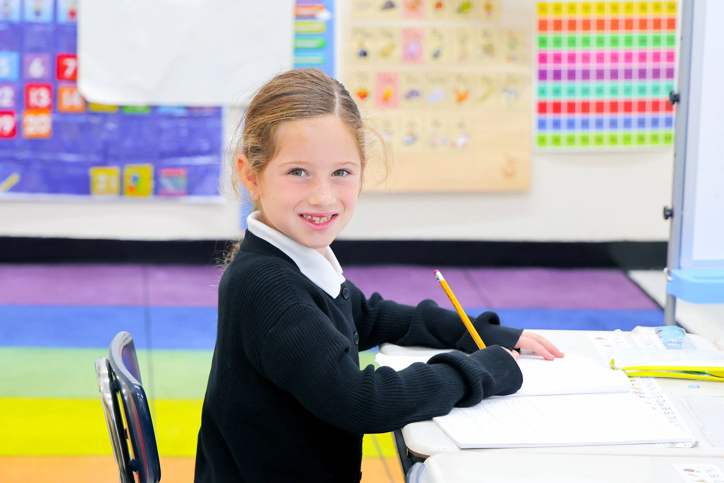 A young girl with light brown hair and freckles sitting at a desk in a classroom, smiling, with colorful educational posters in the background.