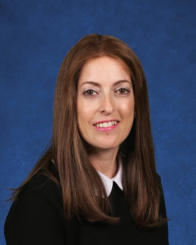 A woman with long brown hair wearing a black blazer and a white collared shirt, smiling, with a blue background.