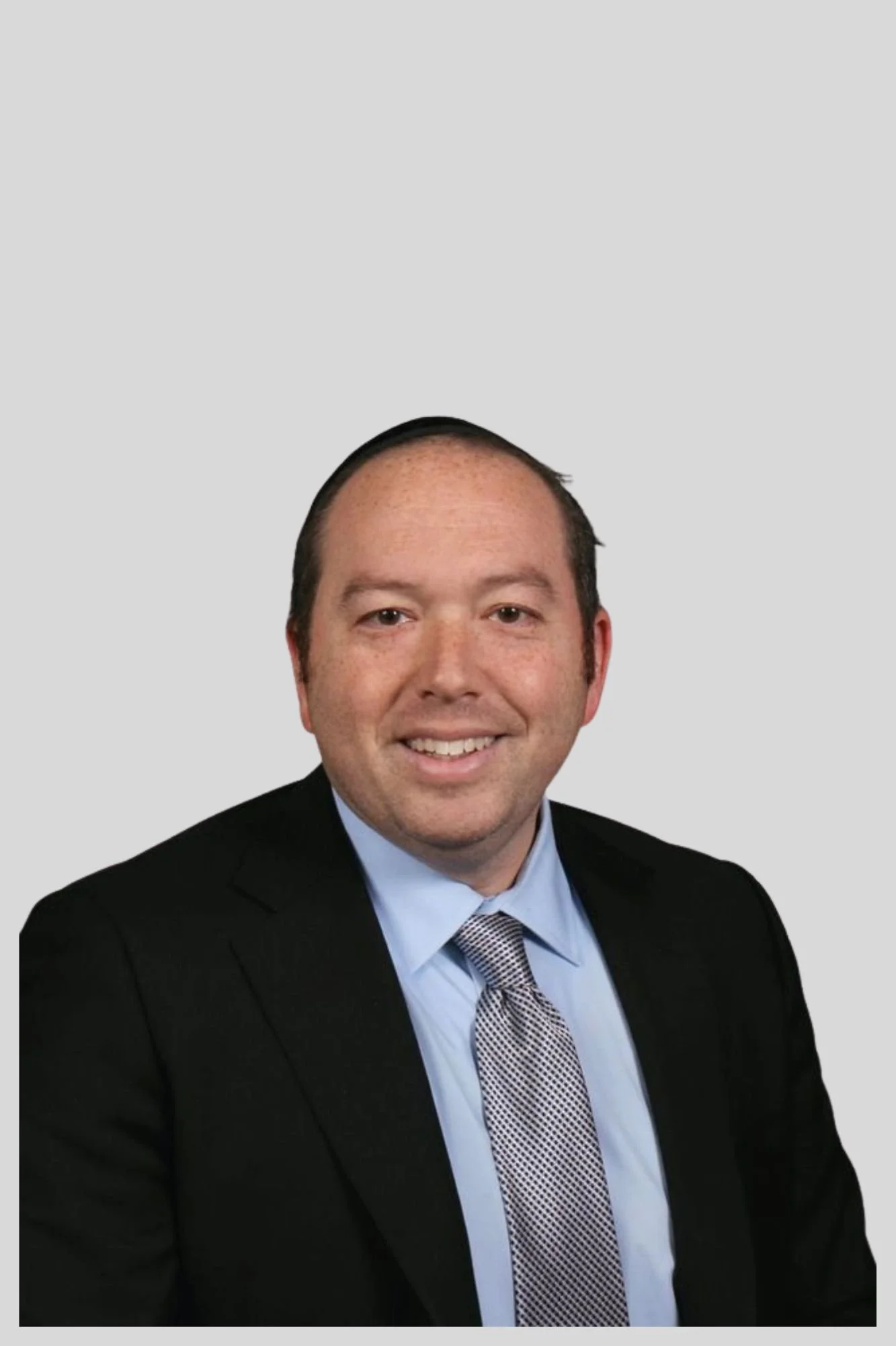 Headshot of a man wearing a black suit, light blue shirt, and patterned gray tie, smiling against a plain light gray background.
