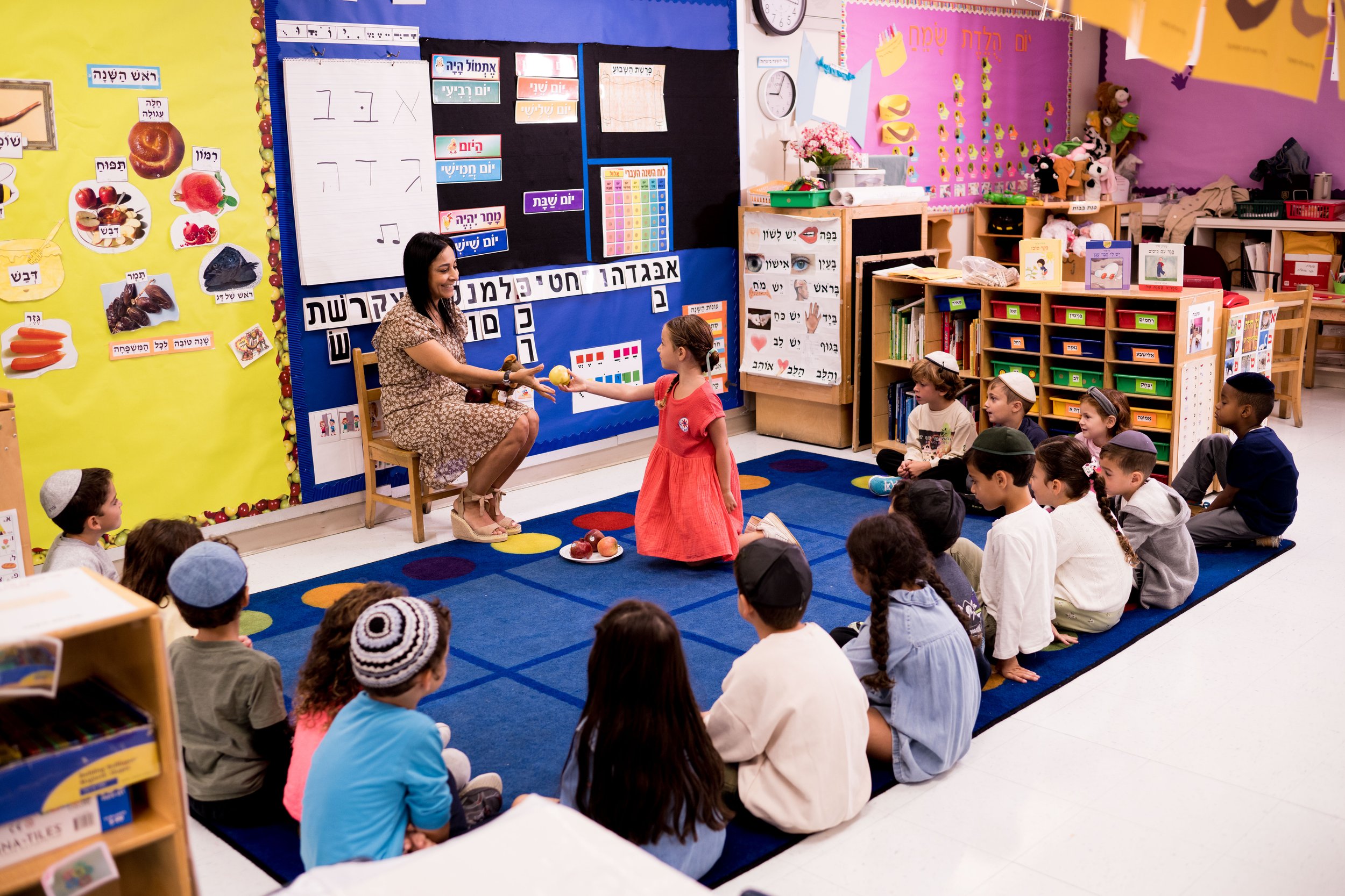 A classroom with young students sitting on a blue and multicolored rug, listening to a teacher who is handing an apple to a girl in a red dress. The classroom has colorful decorations, bookshelves, posters with Hebrew words and pictures, and a blackboard with educational content.
