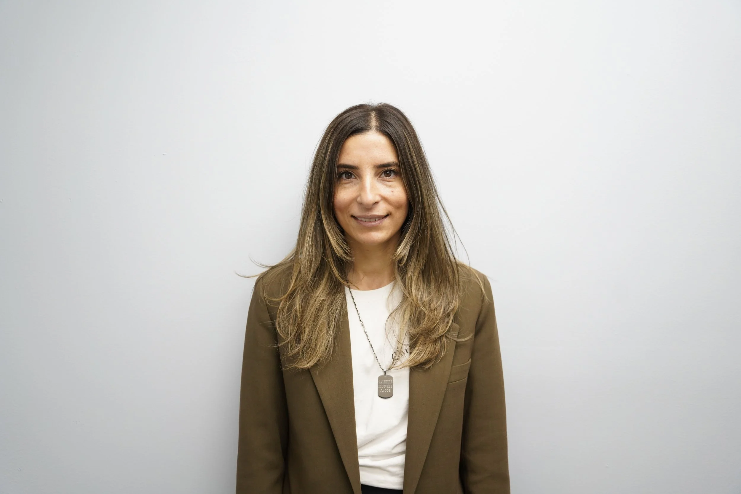 A woman with long brown hair, wearing a brown blazer over a white shirt, standing against a plain light gray wall.