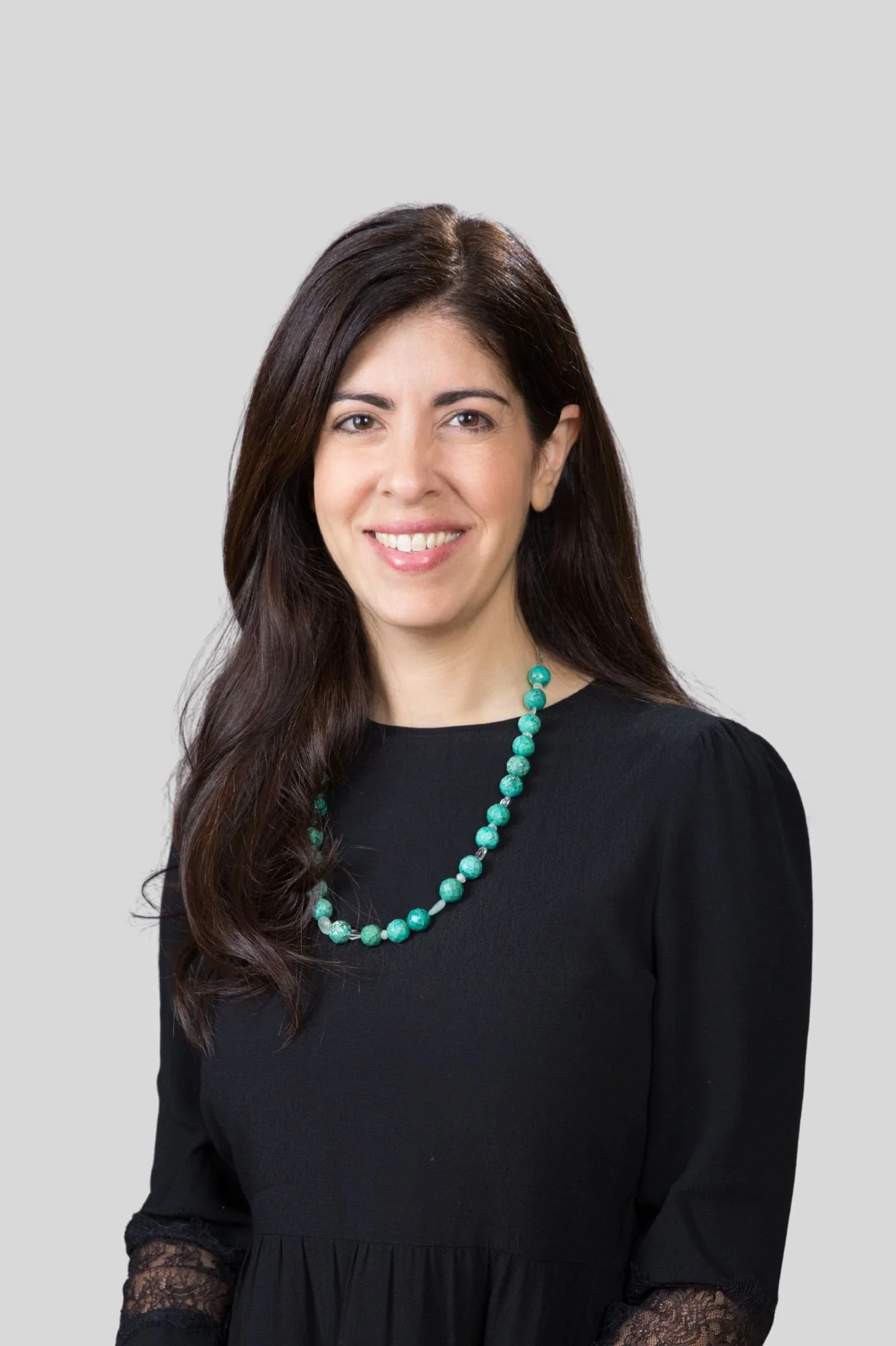 A woman with long brown hair wearing a black dress with lace sleeves and a turquoise bead necklace, smiling at the camera against a light gray background.