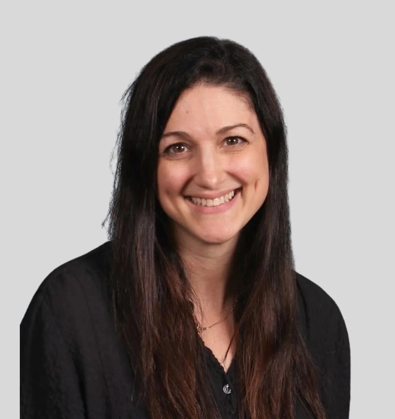 A woman with long dark brown hair, smiling, wearing a black top and a necklace, against a light grey background.