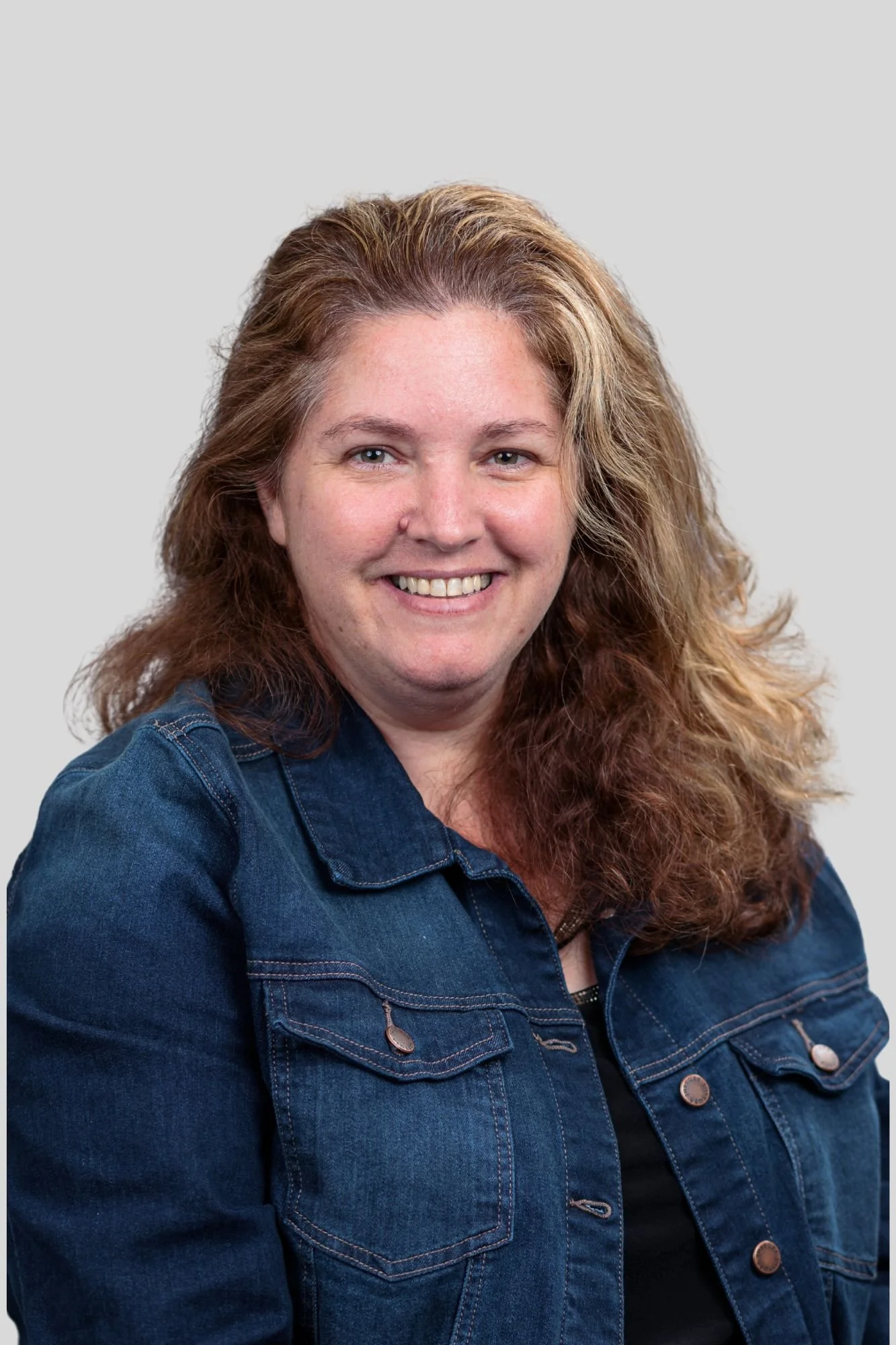 Portrait of a woman with long, curly red hair smiling, wearing a denim jacket and black top against a plain light gray background.