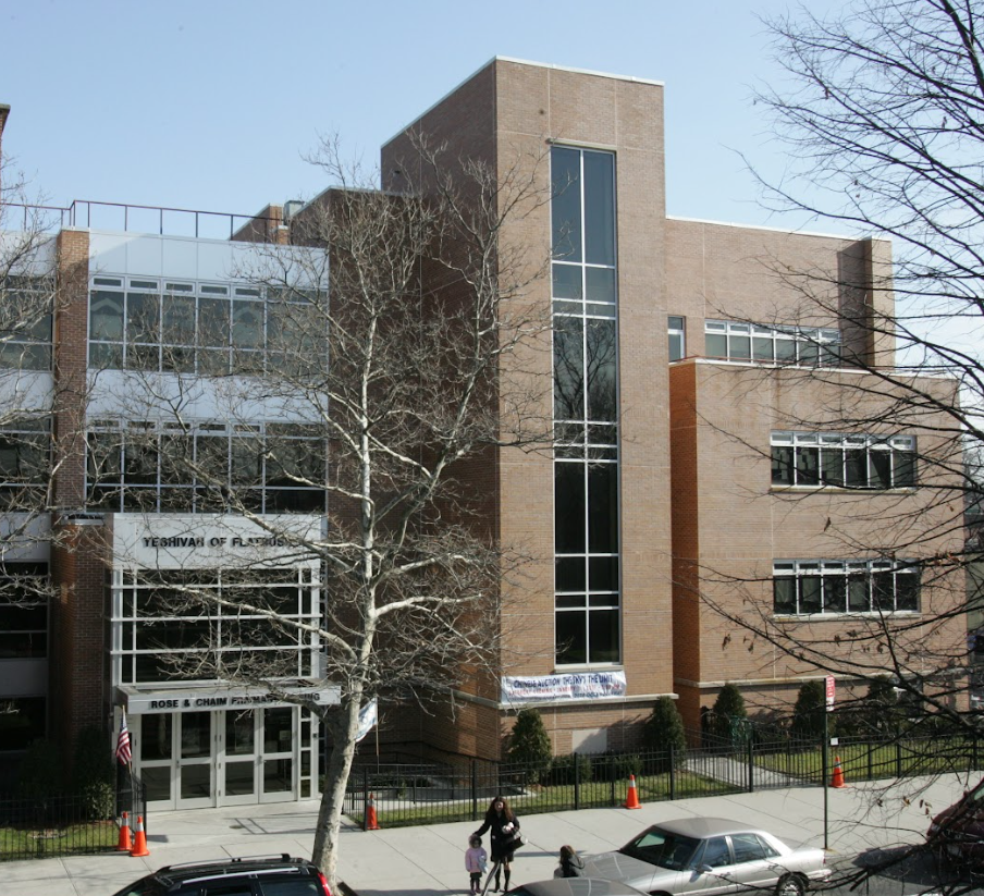 Multistory educational building with brick and glass exterior, trees in foreground, cars parked in front, and people walking near entrance.