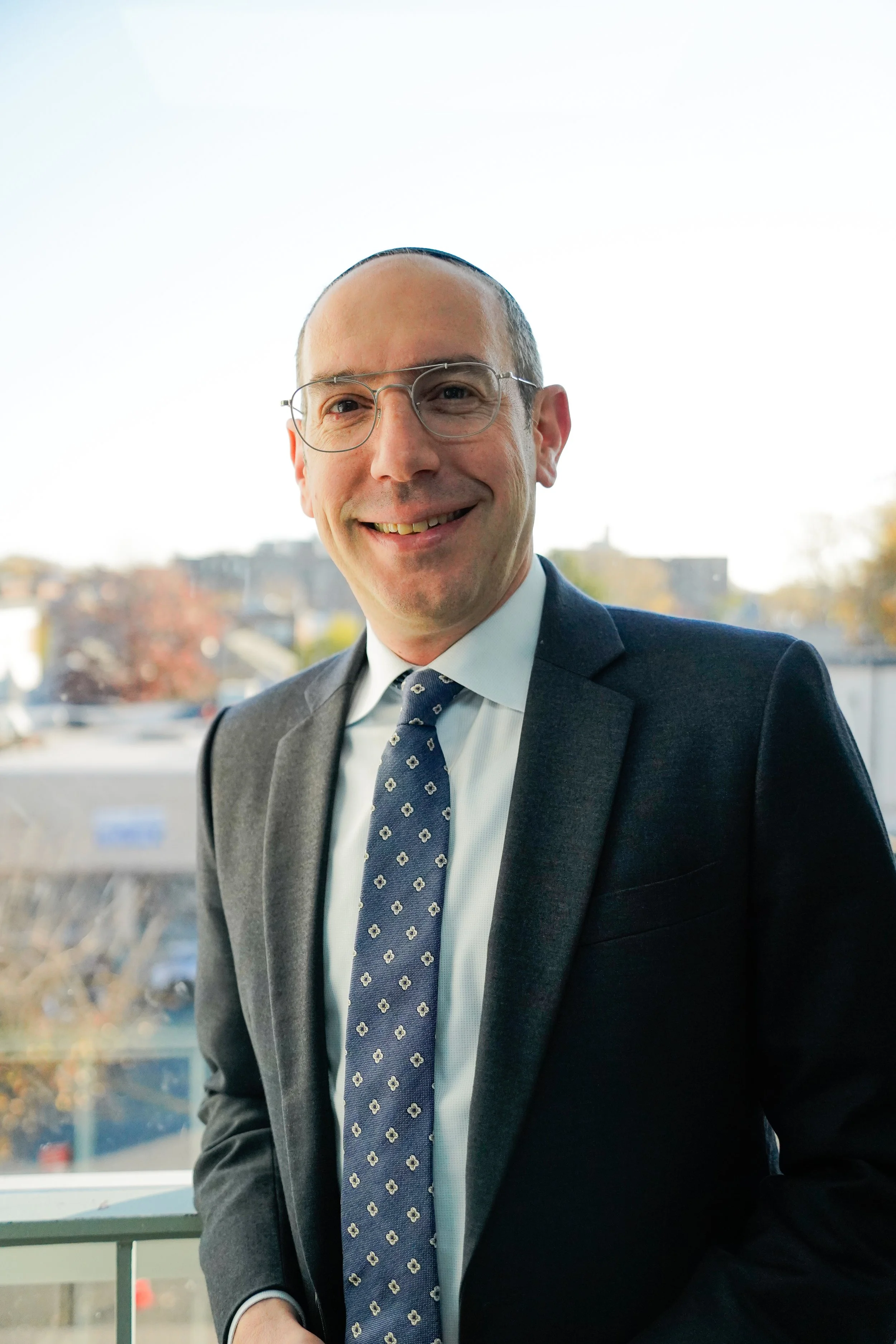 A man in a dark suit, light blue shirt, and patterned tie smiling indoors near a window with a cityscape in the background.