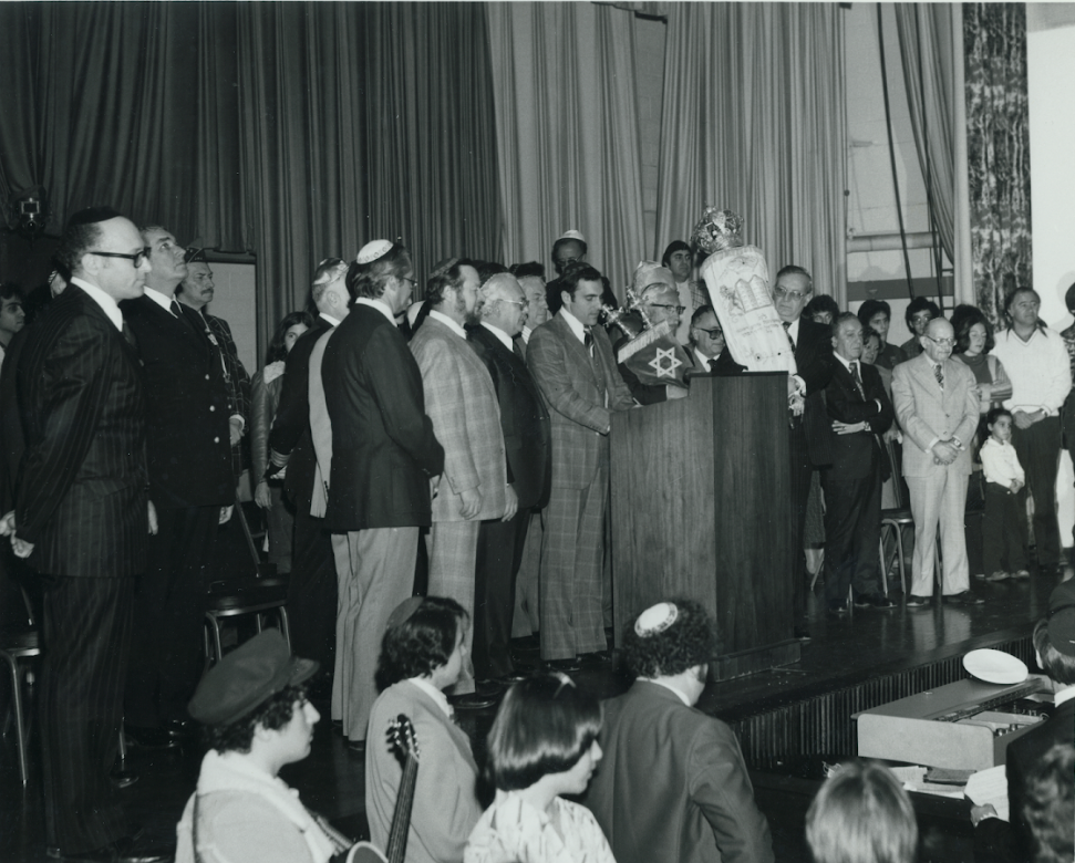 A black and white photo of a group of men and women on a stage, some wearing kippahs, with one man playing a clarinet and a few others holding microphones and standing behind a lectern. The audience in the foreground includes women with a guitar and a man with a yarmulke.