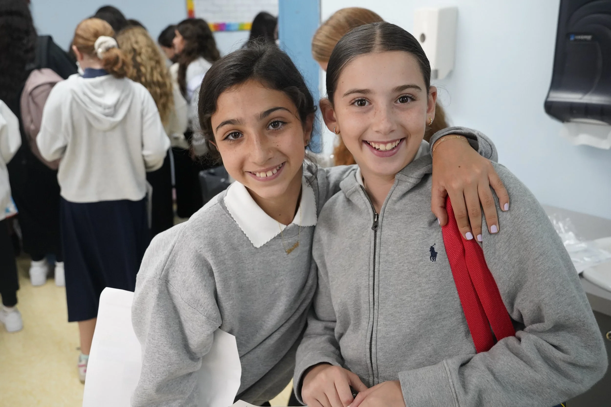 Two young girls smiling and hugging in a classroom, with other children blurred in the background.