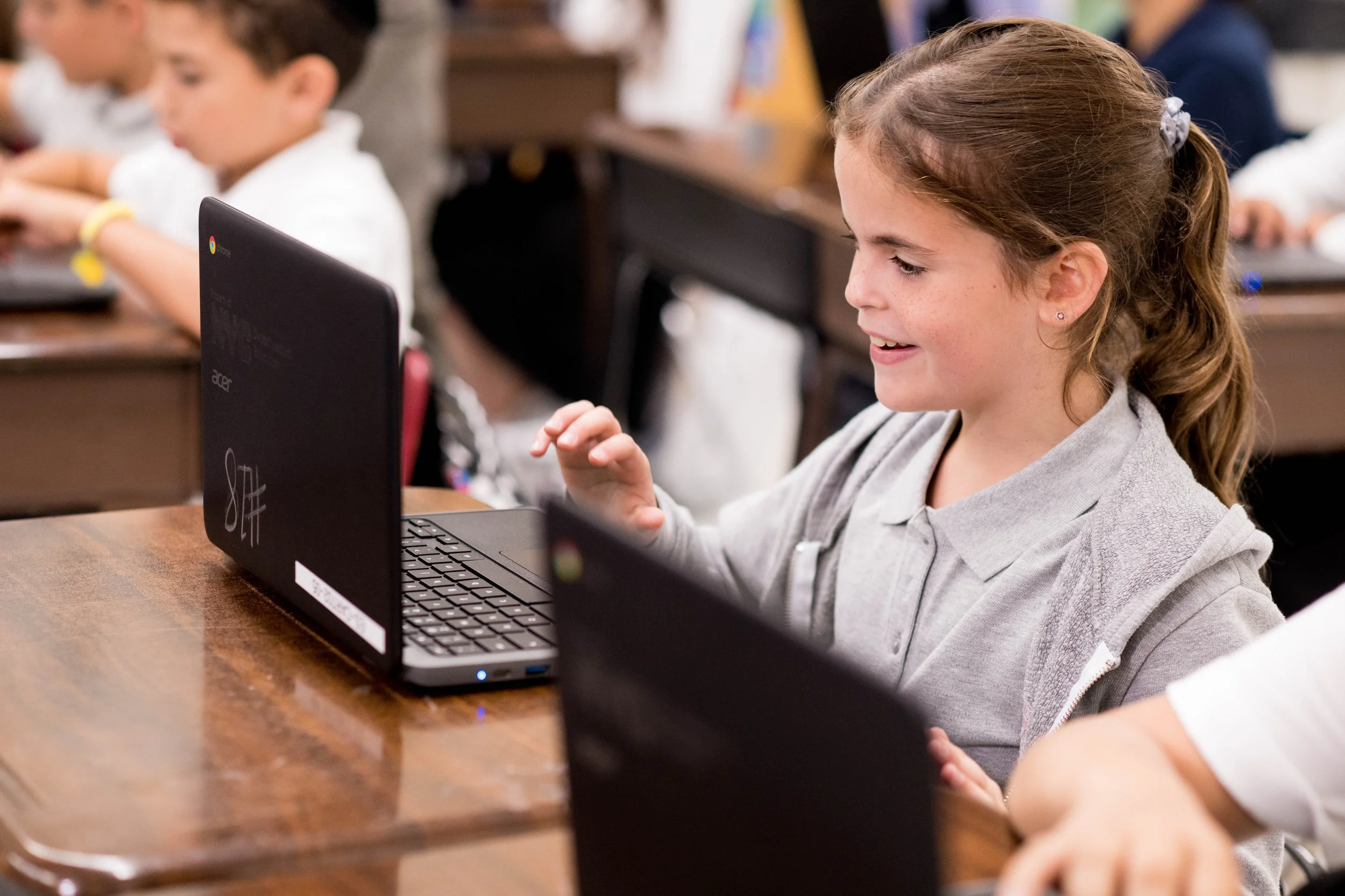 A young girl with brown hair tied in a ponytail, sitting at a wooden desk, working on a laptop in a classroom with other students in the background.