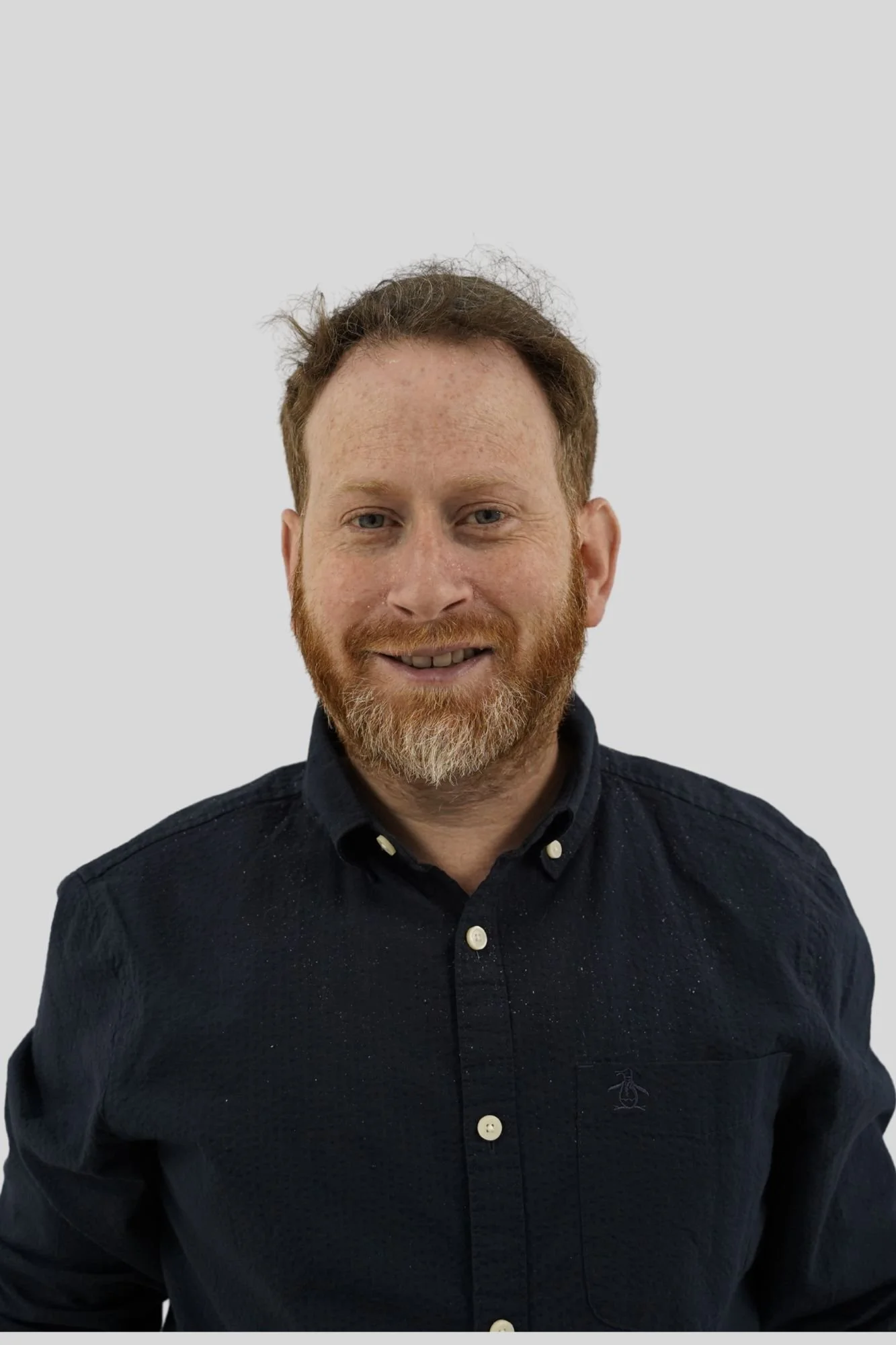 Headshot of a man with a beard, reddish hair, blue eyes, wearing a dark button-up shirt, smiling in front of a light gray background.