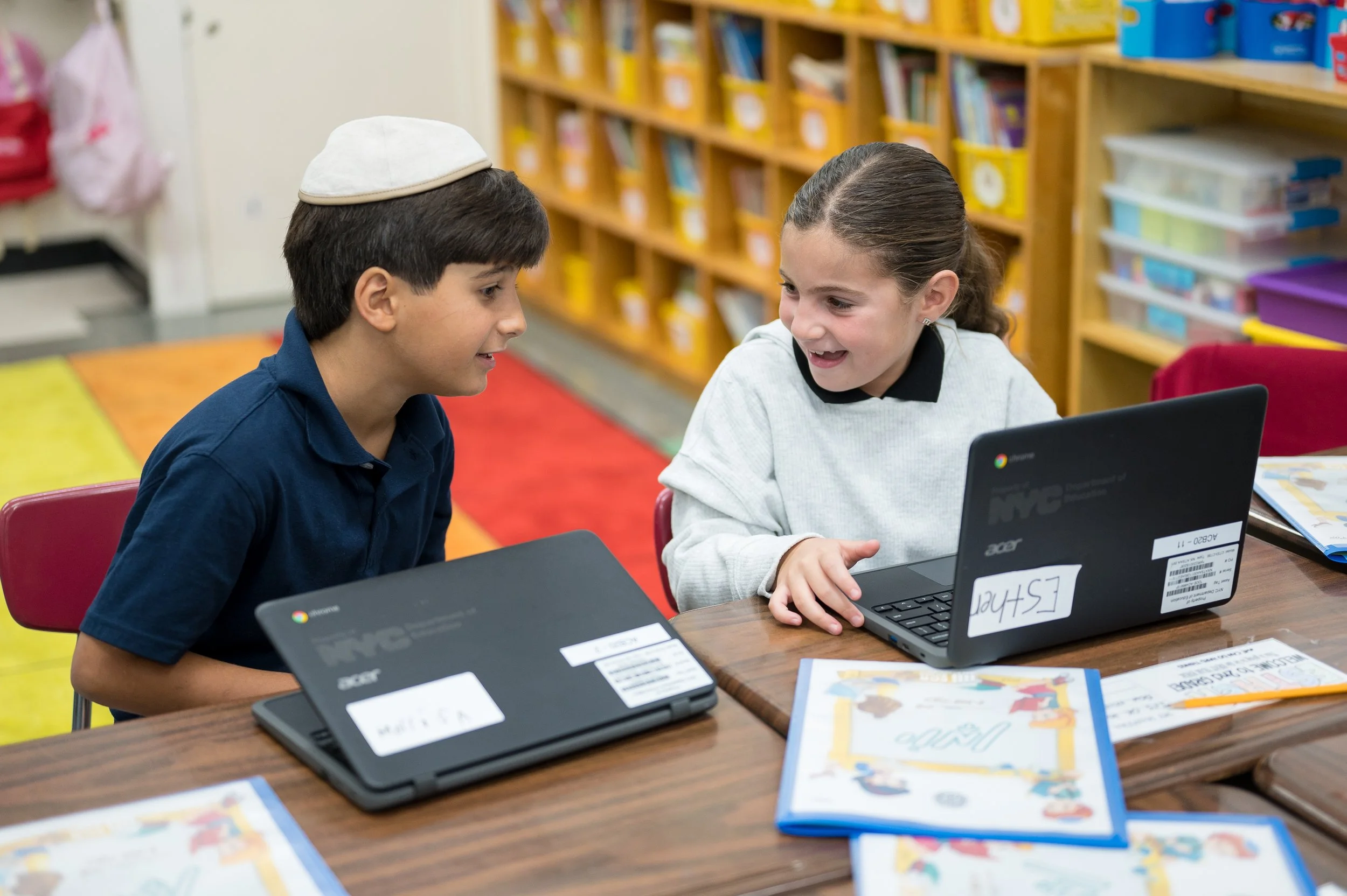 A young boy and girl sitting at a classroom table, smiling and engaging with each other. The boy is wearing a blue shirt and a yarmulke, and the girl is in a white top. There are laptops, books, and colorful educational materials on the table.