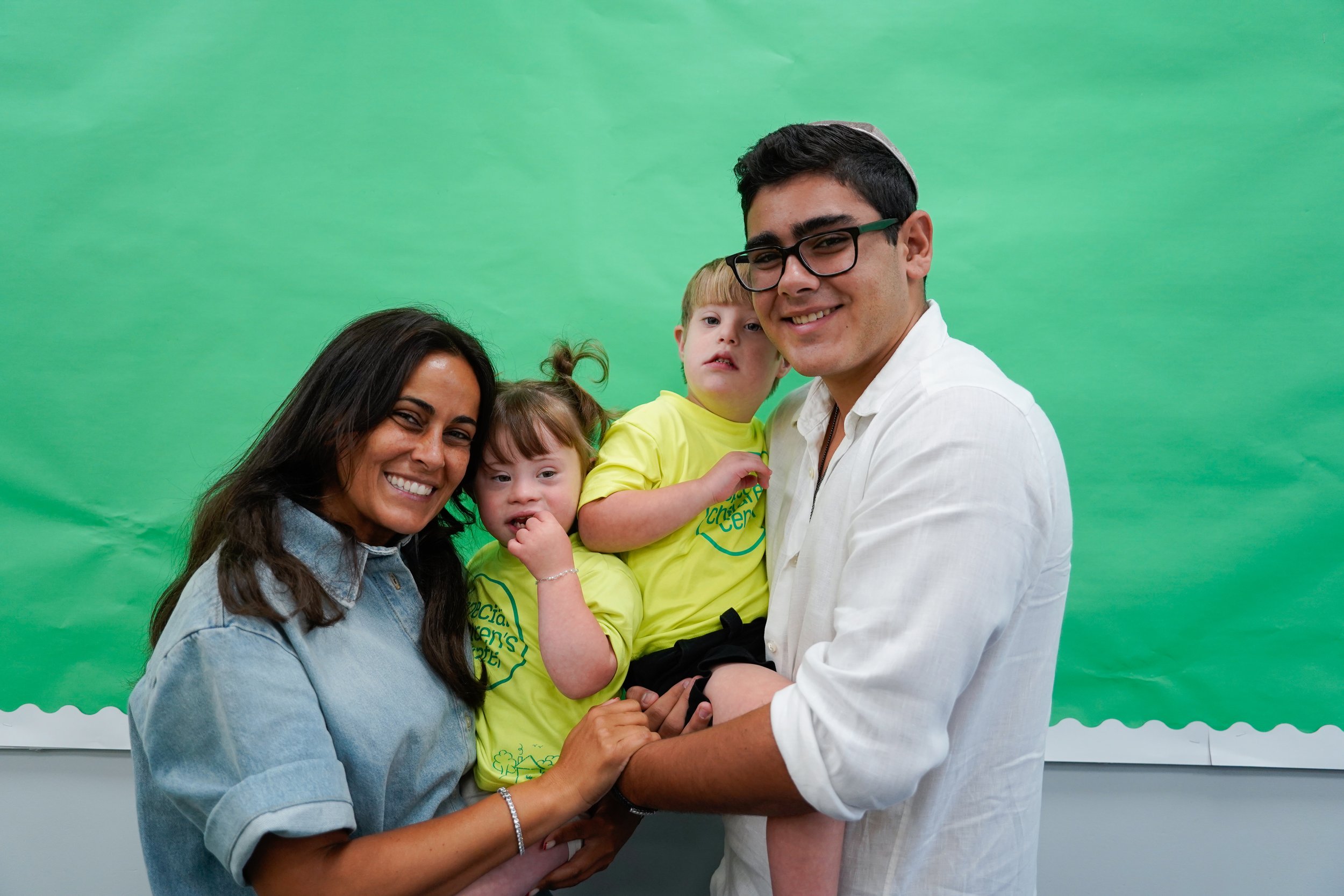 A smiling family of four holding hands in front of a green backdrop.