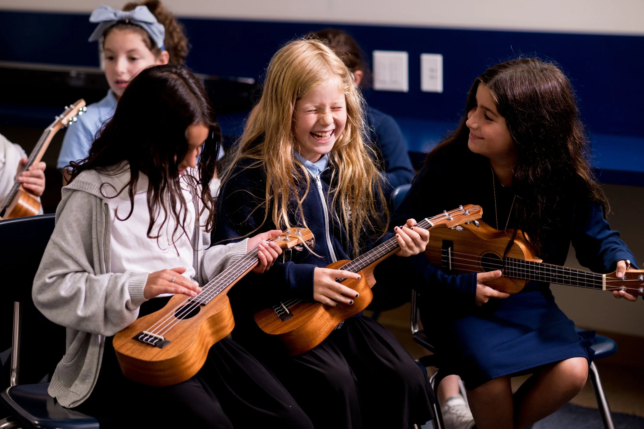 Group of young girls sitting in a music classroom, playing ukuleles, smiling, and enjoying a music lesson.