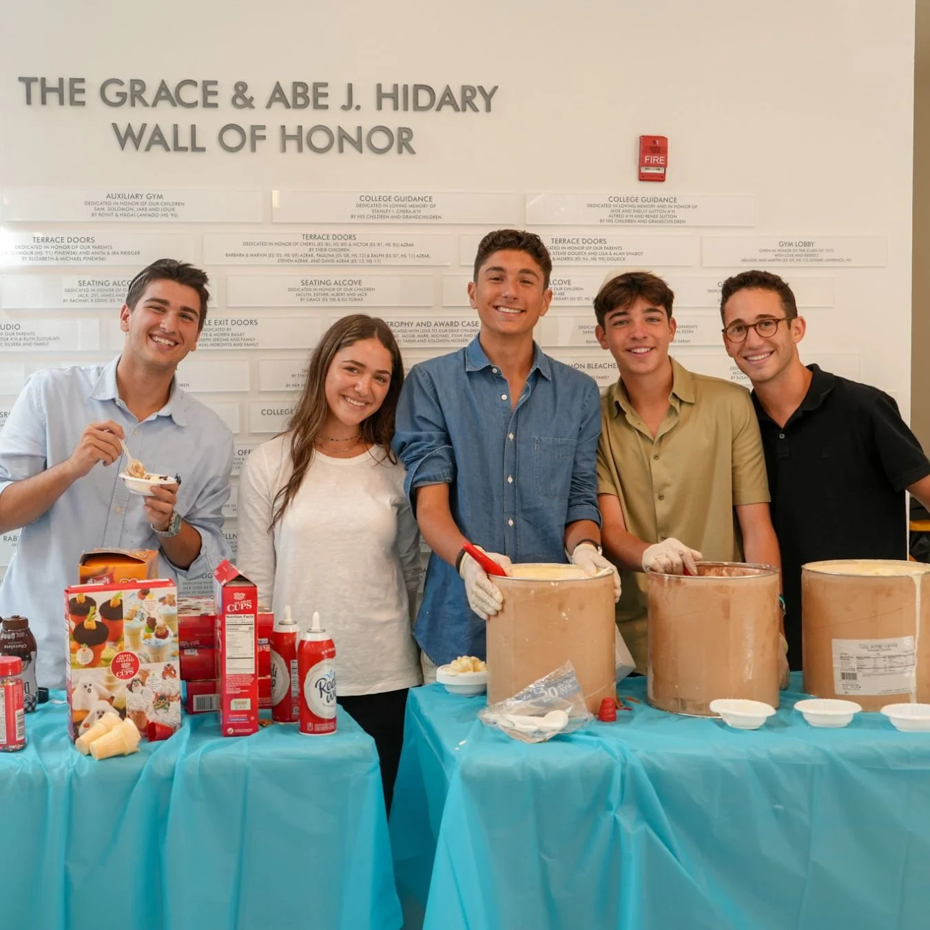 A group of five young people standing at a table with ice cream supplies in front of a wall with the text 'The Grace & Abe J. Hidy Wall of Honor', smiling and enjoying a communal activity.