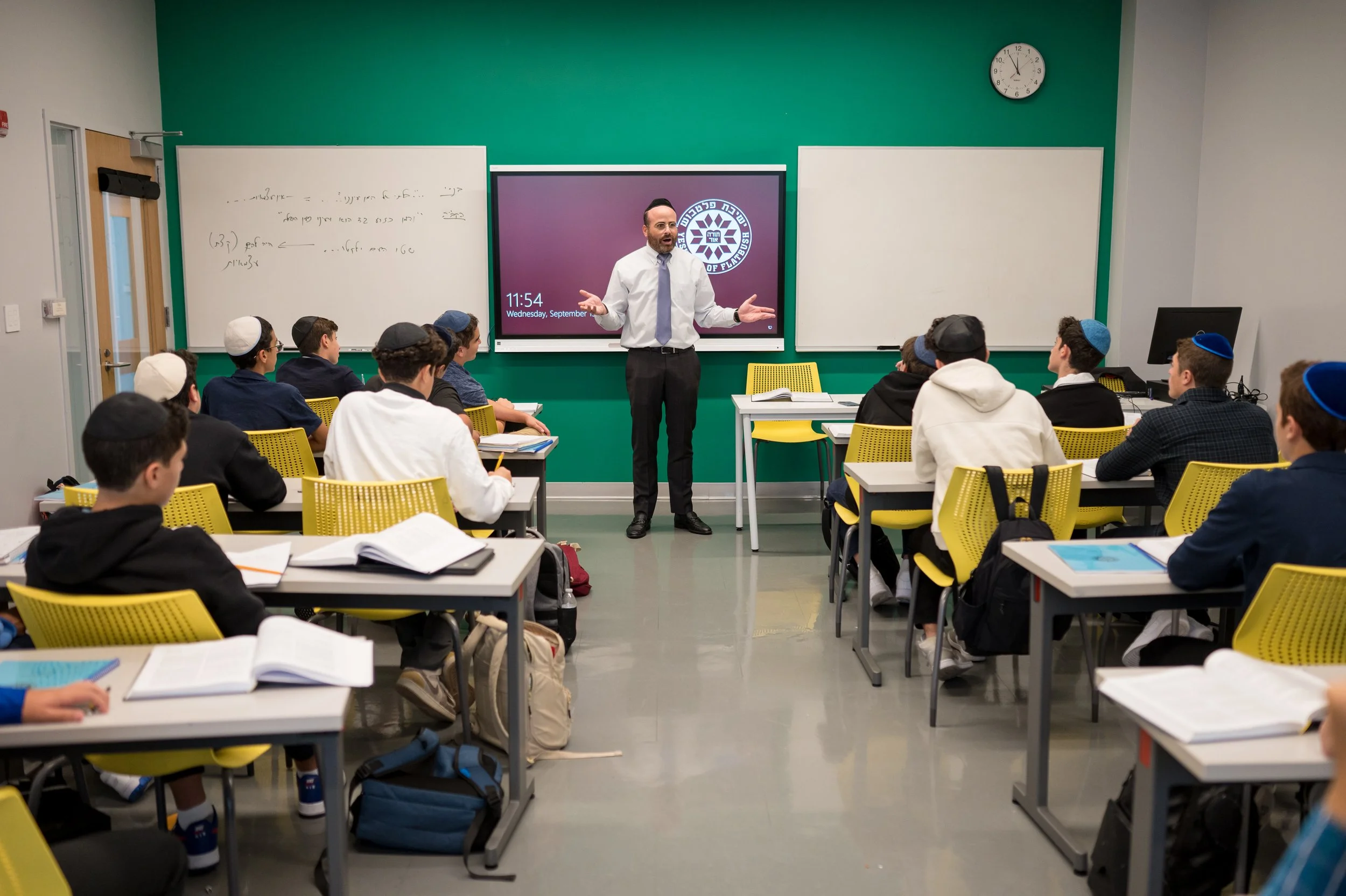 A classroom with students sitting at desks and a teacher standing at the front gesturing with his hands. The classroom has green and beige walls, a digital clock showing 11:54, whiteboards, and a screen with a logo. The students are wearing kippahs a