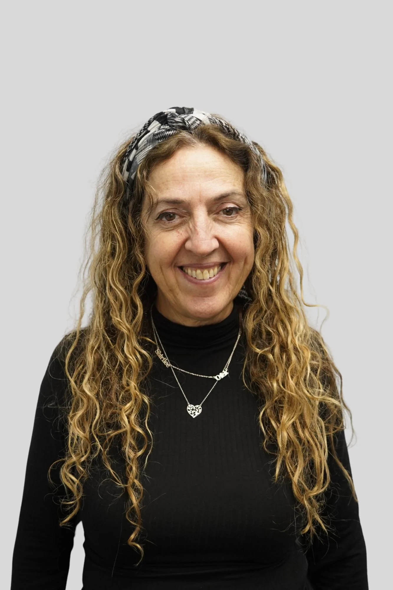 Portrait of a woman with long curly hair wearing a black turtleneck and decorative jewelry, including heart-shaped necklace and layered chain necklaces, smiling at the camera against a plain light grey background.