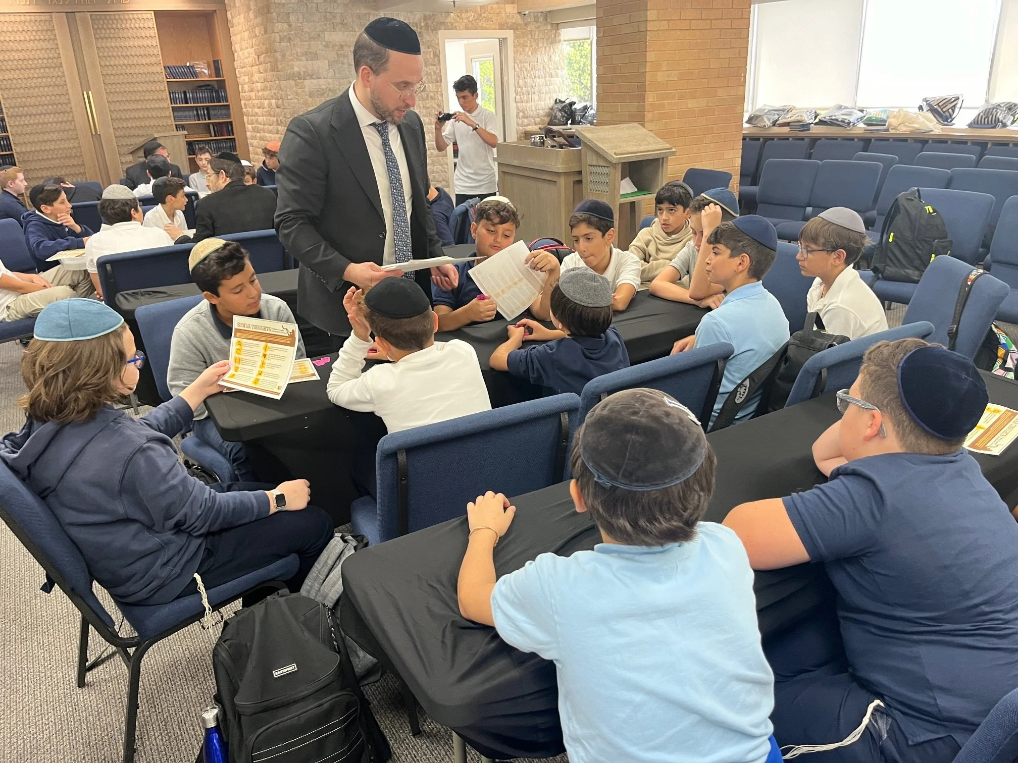 A group of young boys and a man, likely a teacher or rabbi, sitting around a table in a classroom or study hall. The man is standing and reading from a book, and several boys are wearing kippahs. The room has shelves of books, backpacks, and windows bringing in natural light.