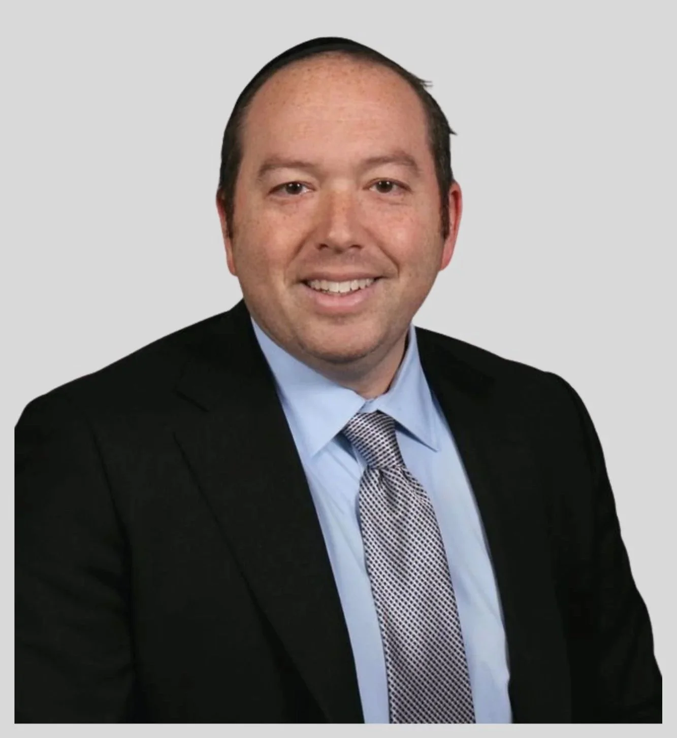 Headshot of a man in a suit with a blue shirt and patterned tie, smiling, against a light grey background.