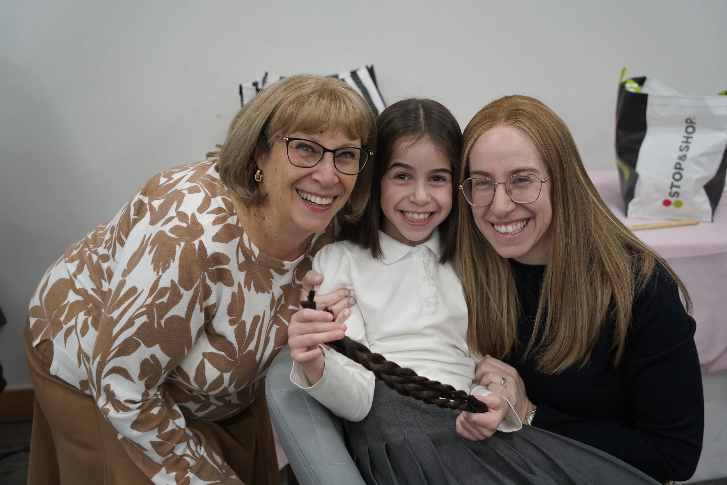 Three females, two older women and a young girl, smiling and posing together for a photo. The women are wearing glasses and both have reddish hair, while the girl has dark, straight hair.