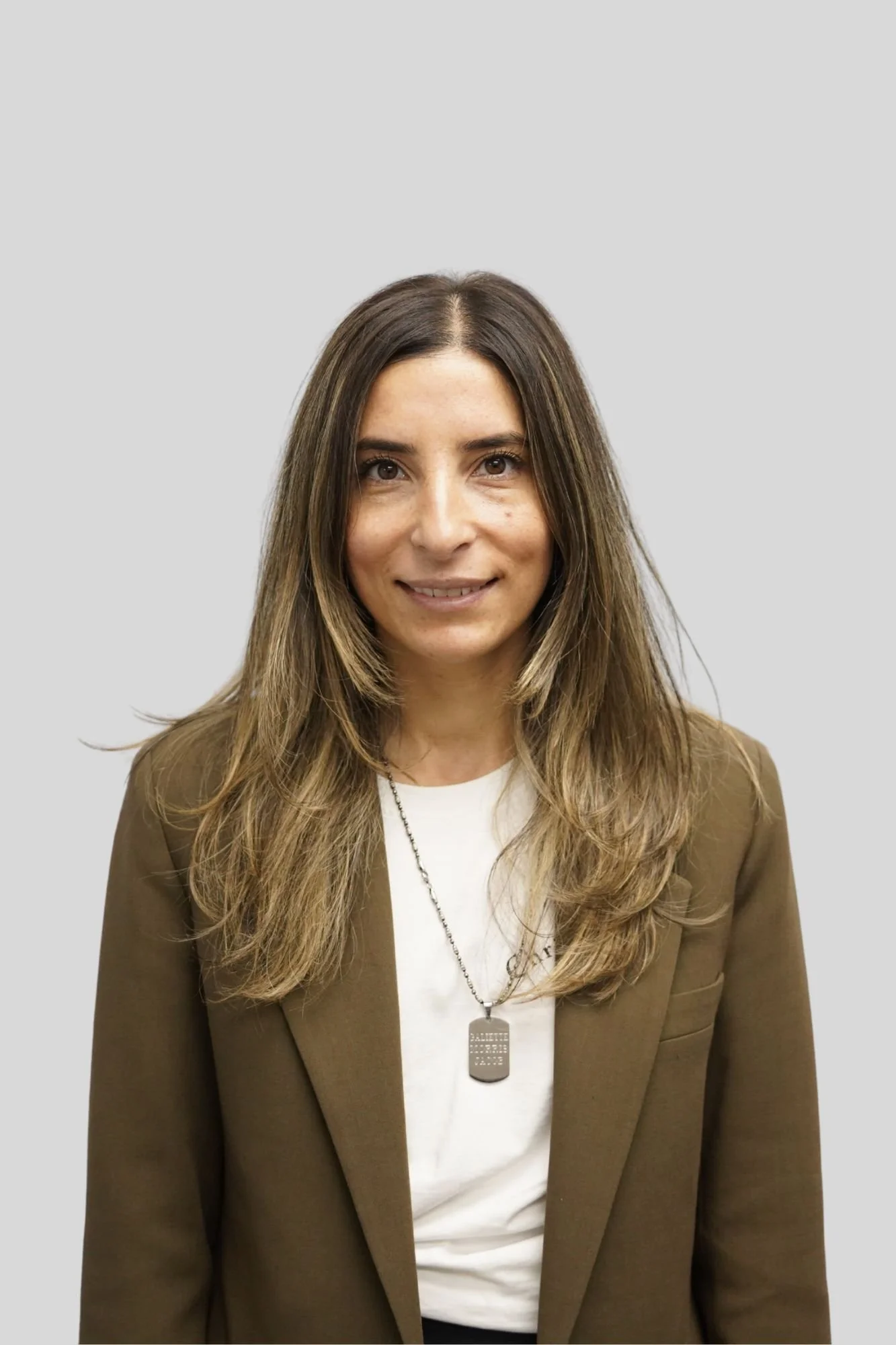 A woman with long, wavy brown hair wearing a brown blazer over a white shirt, with a silver necklace and a dog tag-style lanyard.