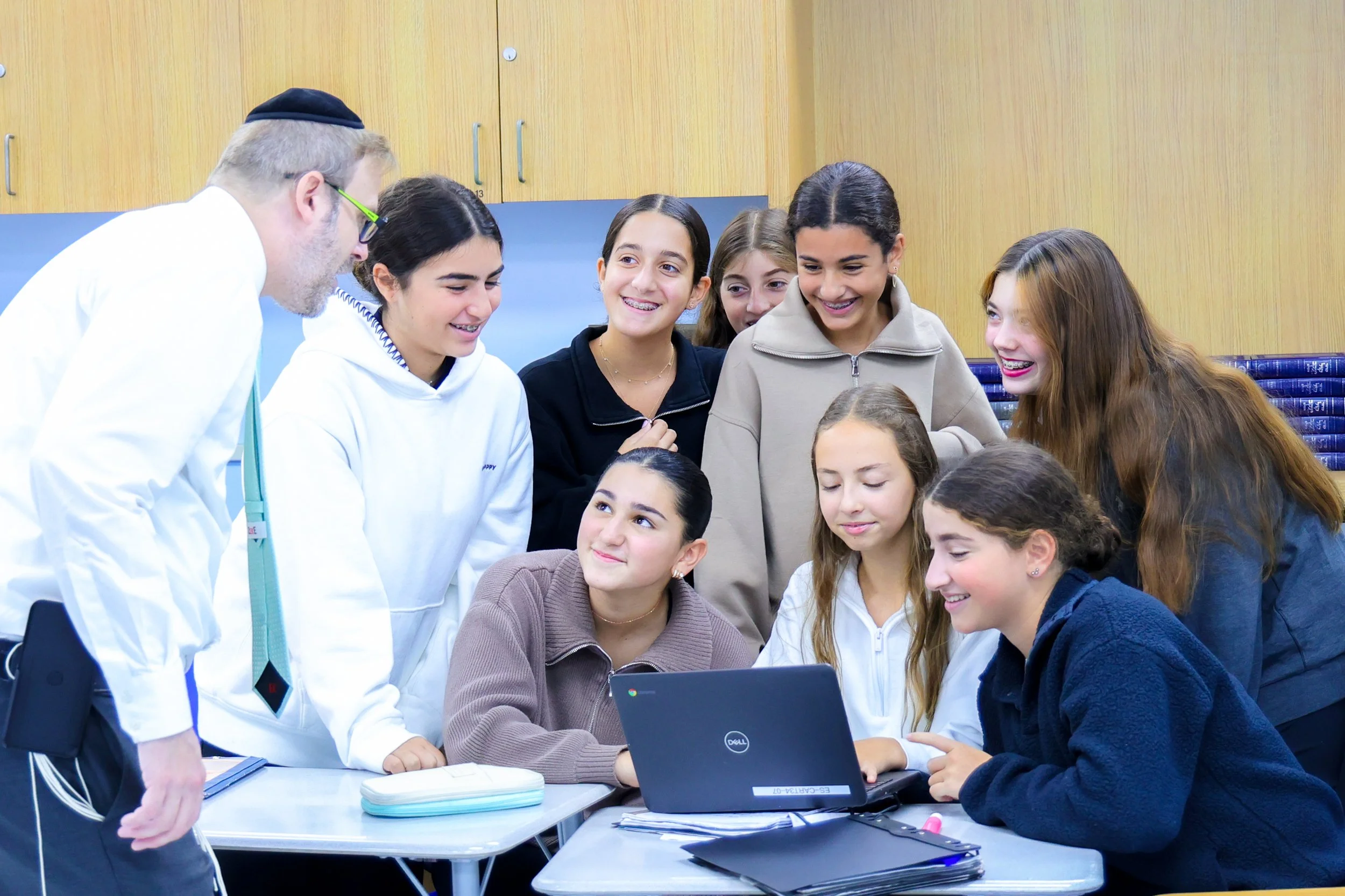 Group of children and a man gathered around a laptop in a classroom.