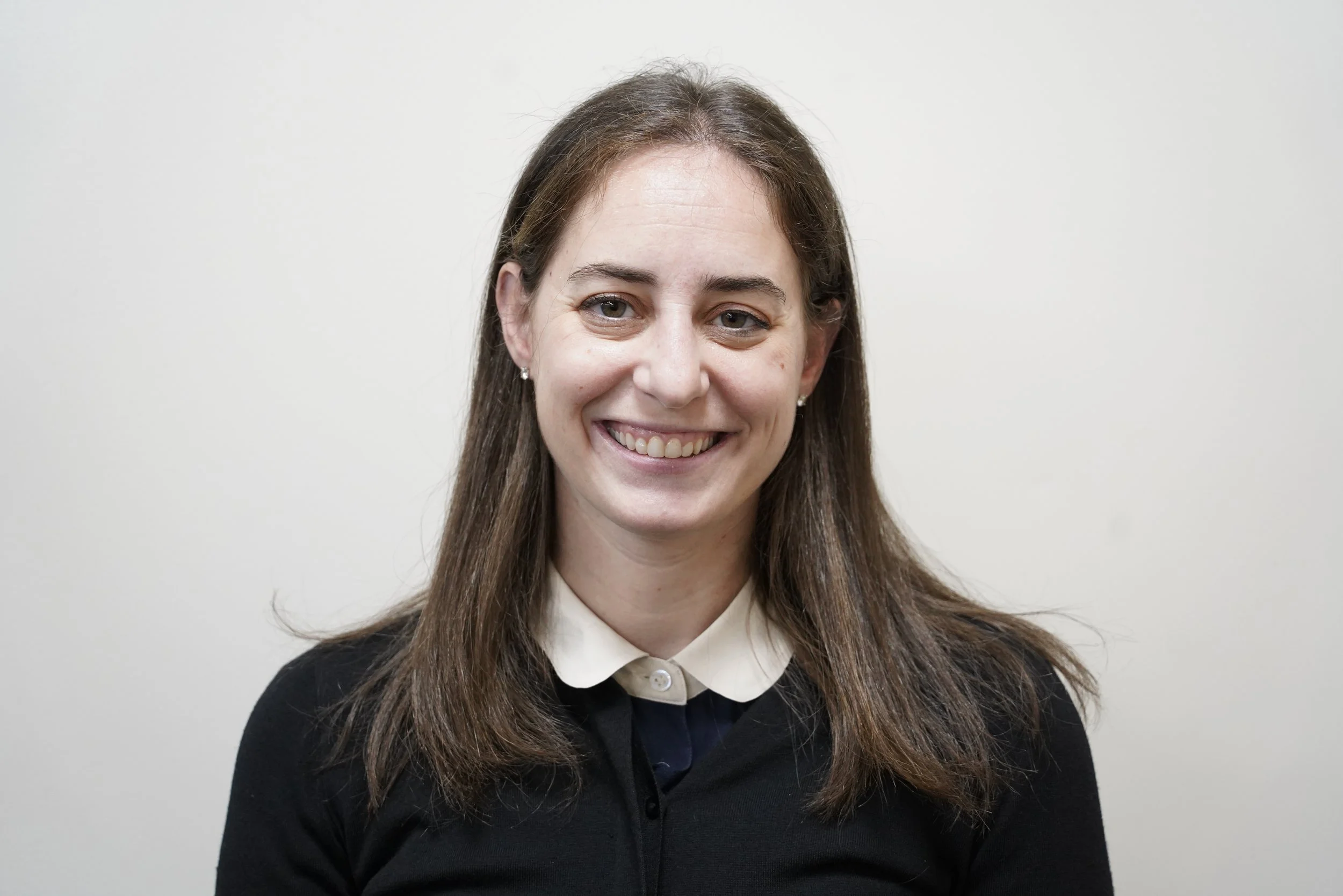 A young woman with long brown hair, wearing a black sweater over a beige collared shirt, smiling against a plain white background.