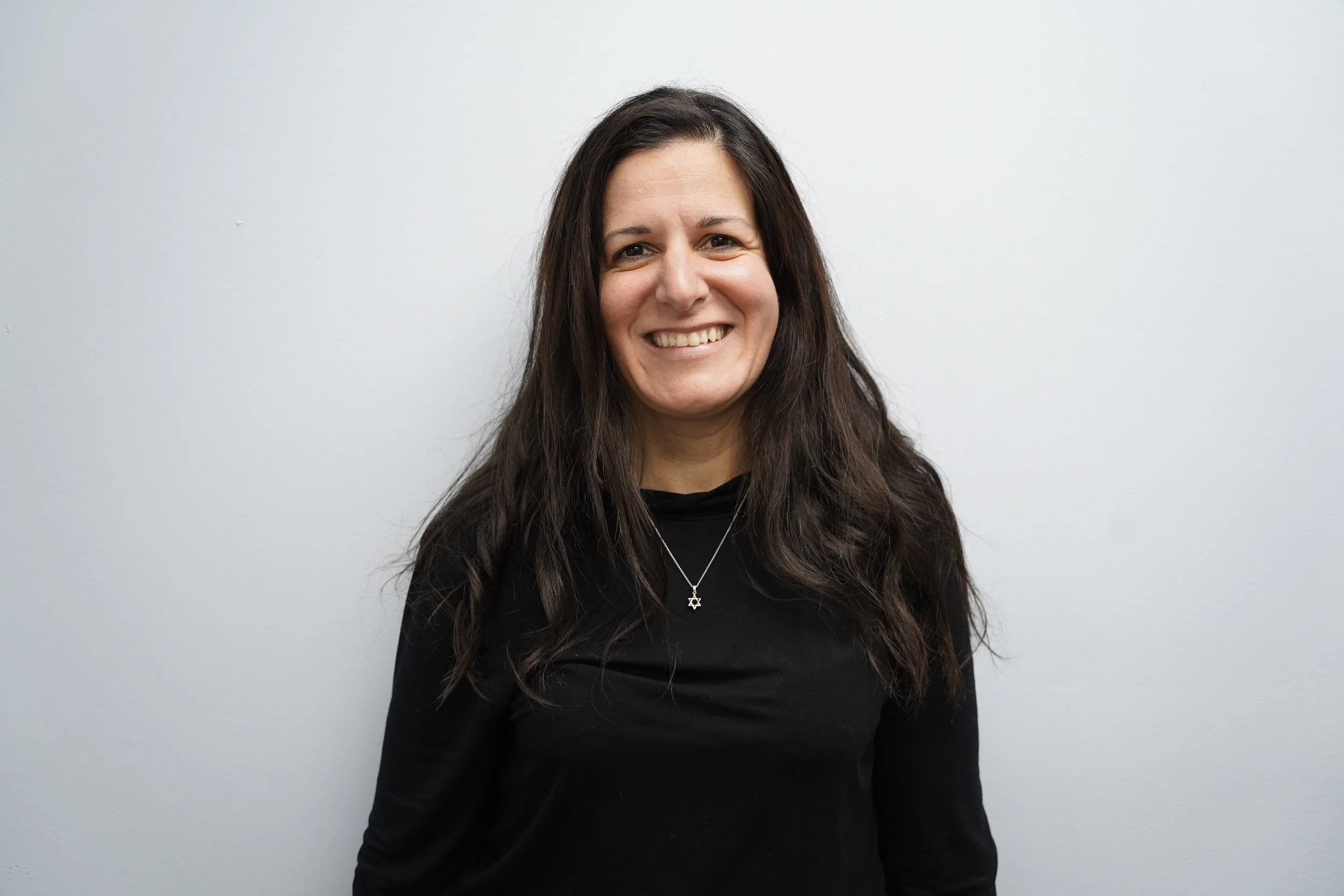 Smiling woman with long dark hair wearing a black top and a Star of David necklace standing against a plain white wall.