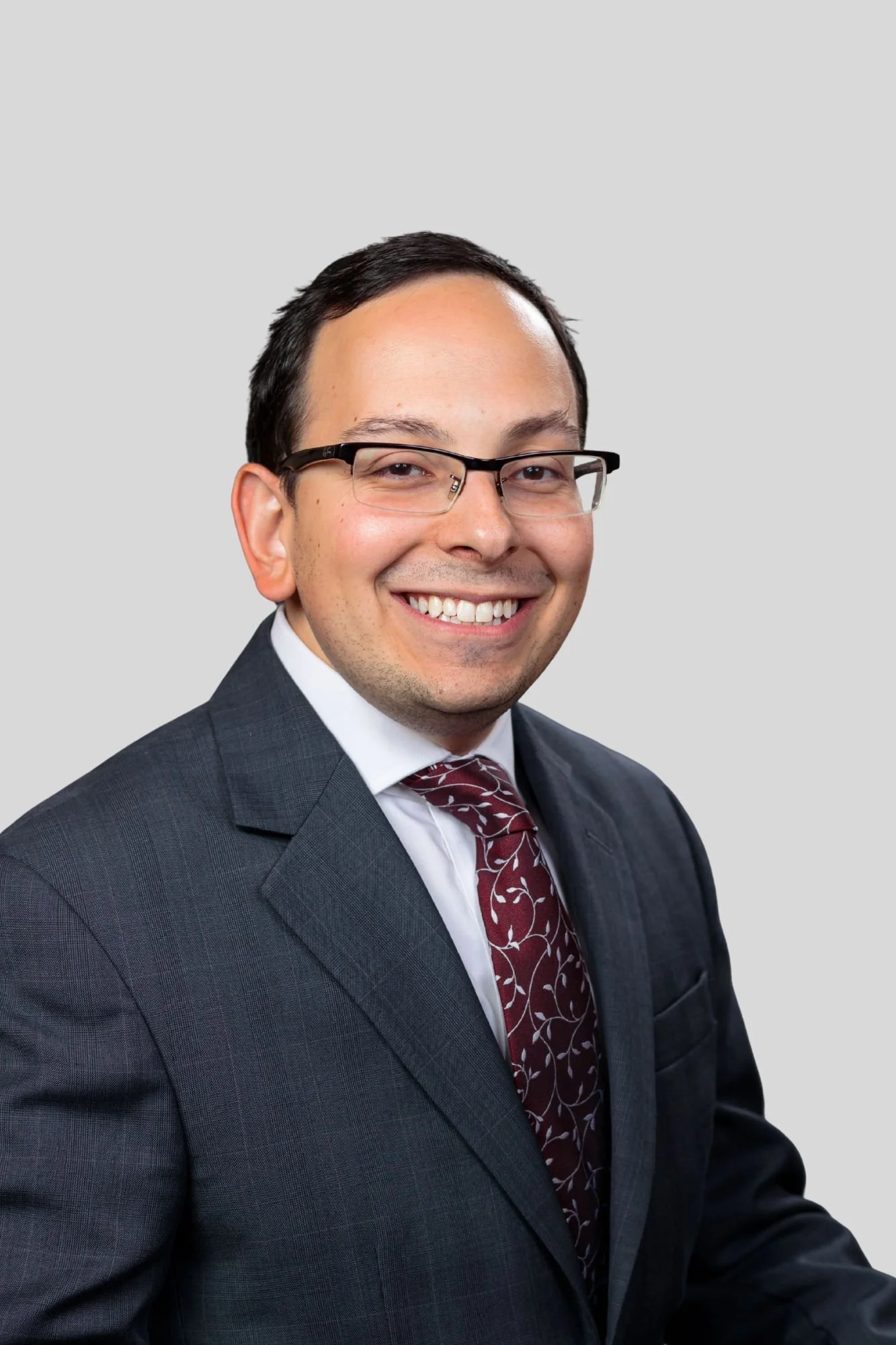 A smiling man dressed in a dark suit, white shirt, and maroon patterned tie, with short dark hair and glasses, posing against a light gray background.