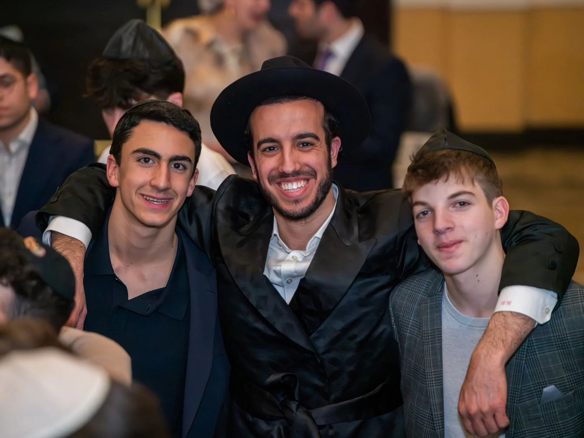 Three young men smiling, with two wearing black hats, standing close together at a social gathering with people in the background.