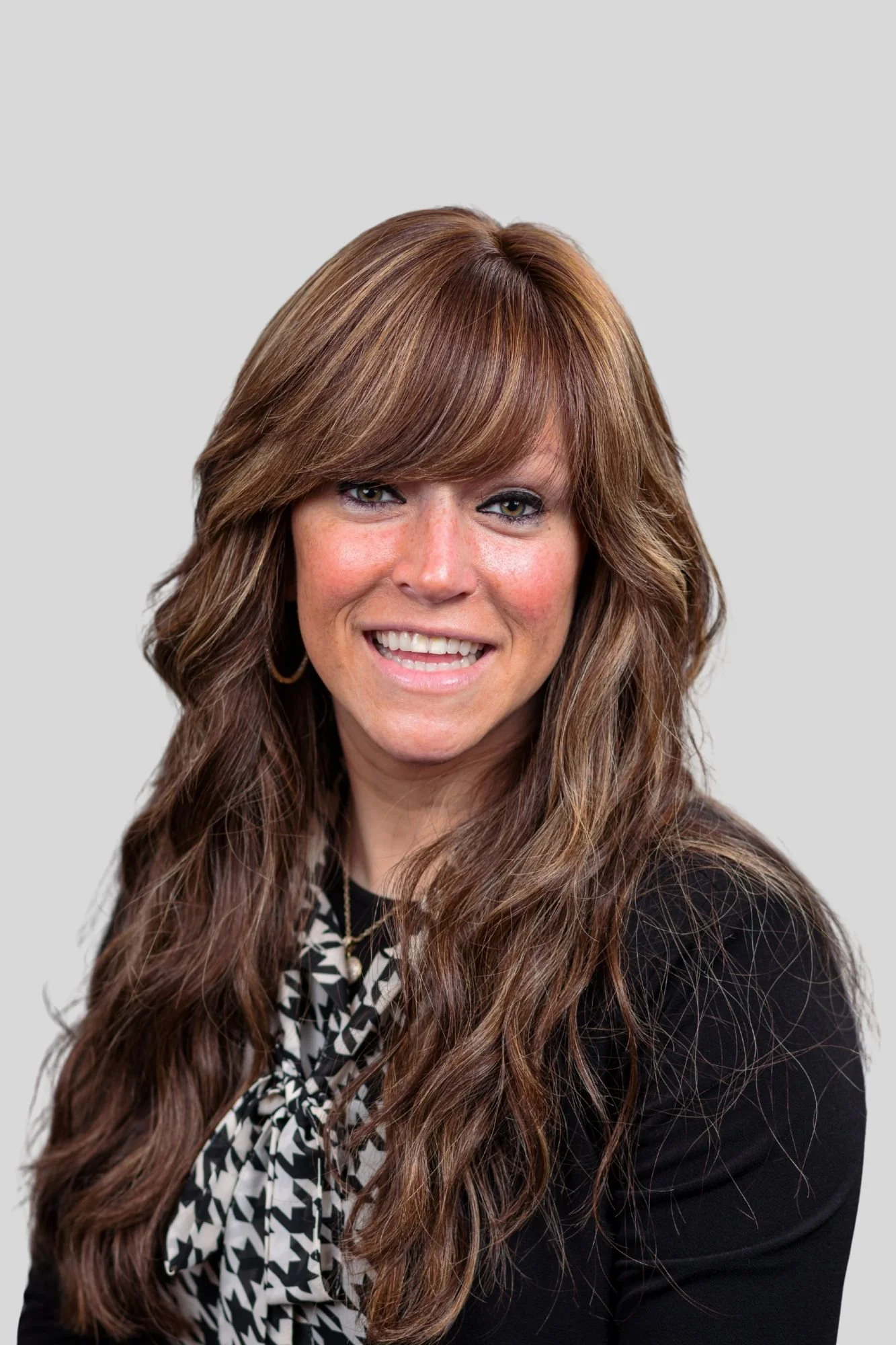 A woman with long, wavy brown hair and light skin, smiling and looking at the camera, wearing a black blazer and a patterned scarf.