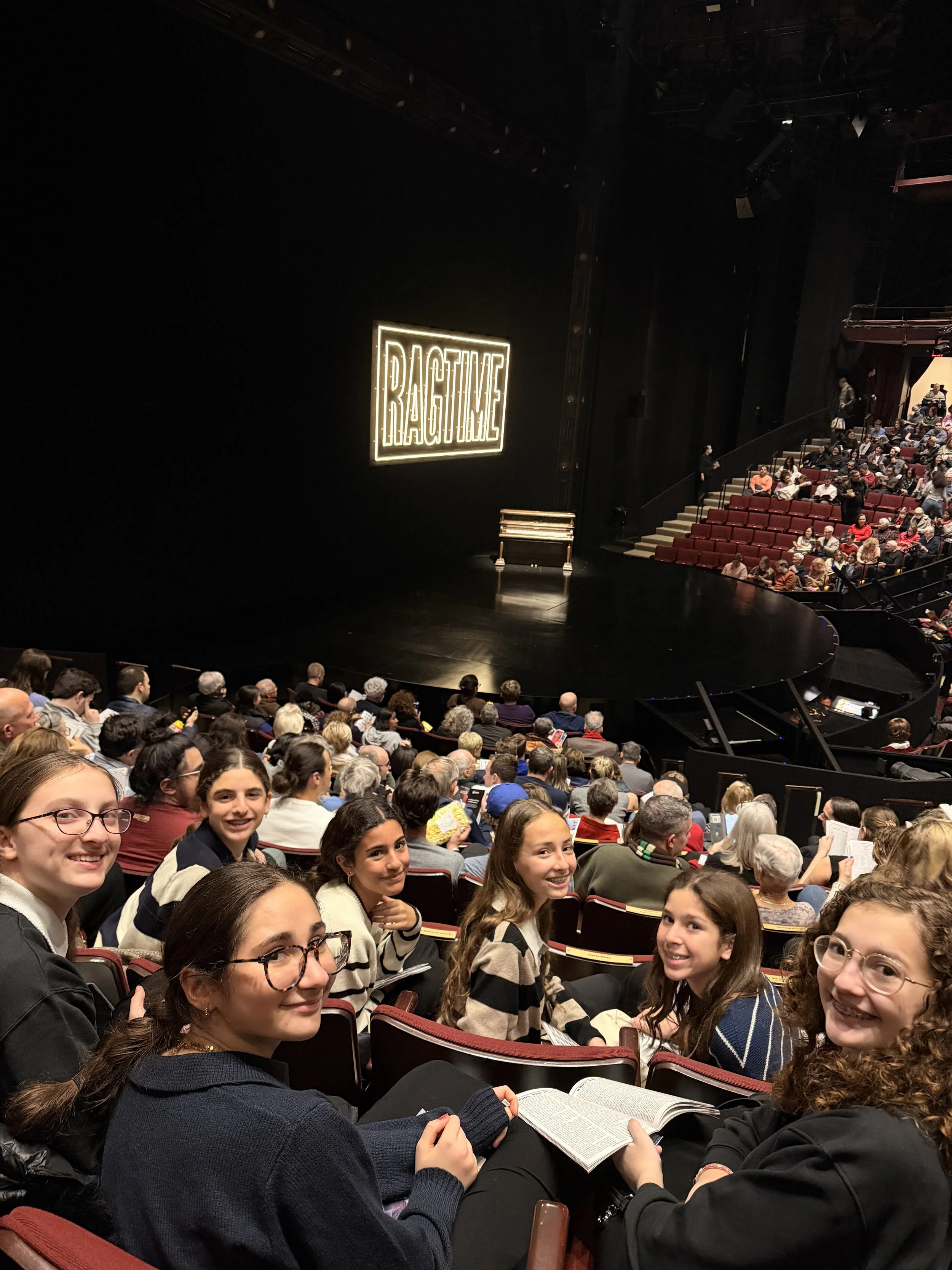A diverse group of young people sitting in theater seats at an event with a stage, some are smiling and talking, with a prominent sign that reads 'RAGTIME' illuminated on the stage.