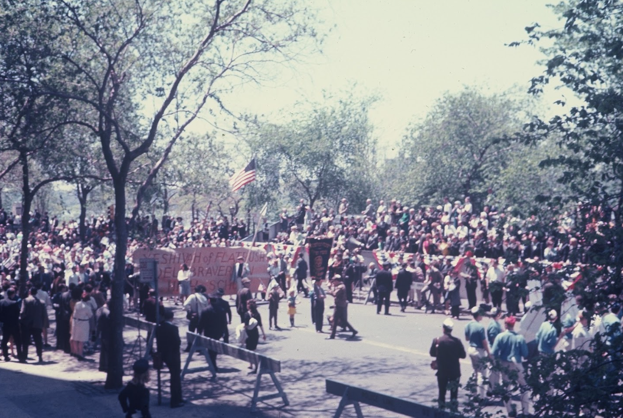 A large group of people gathered outdoors for a parade, with many holding banners and flags. The crowd is watching from the street, with trees and sunlight visible.