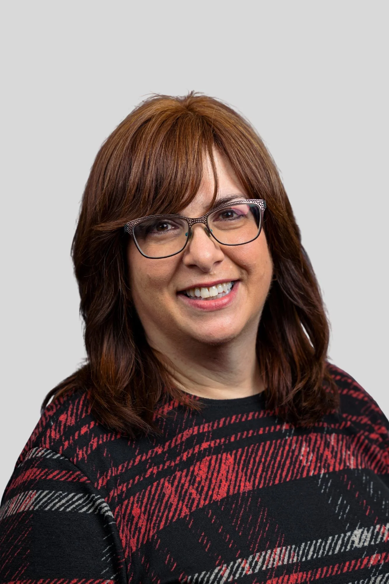 A woman with long, reddish-brown hair wearing glasses and a red and black patterned sweater, smiling at the camera against a plain light gray background.