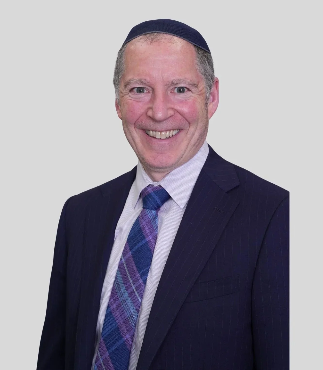 A man in a dark suit with a white shirt and a purple plaid tie, wearing a kippah, smiling against a plain gray background.