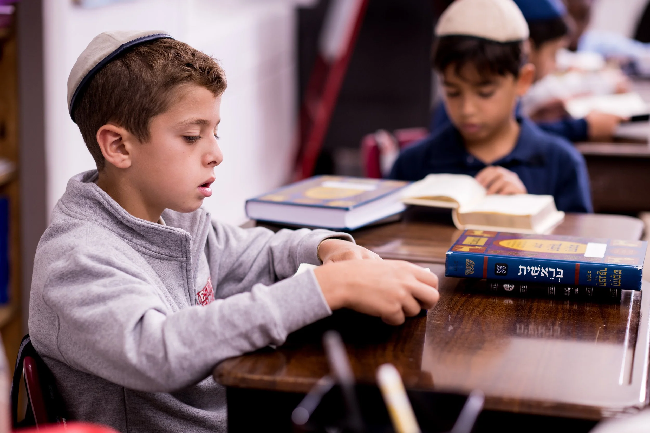 A young boy wearing a kippah, focused on reading a Hebrew Bible at a table in a classroom or study setting.