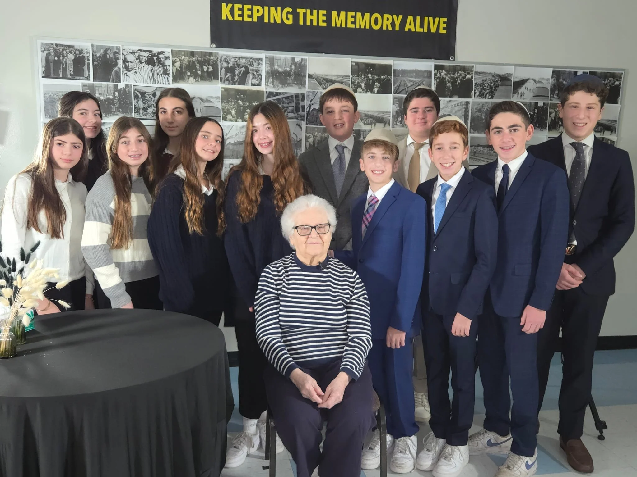 Group of young people dressed in formal attire, standing indoors in front of a black and white historical photographs display. A banner above reads 'Keeping The Memory Alive'. A woman with white hair and glasses seated in the foreground. A black table with a vase of flowers is in front of the group.