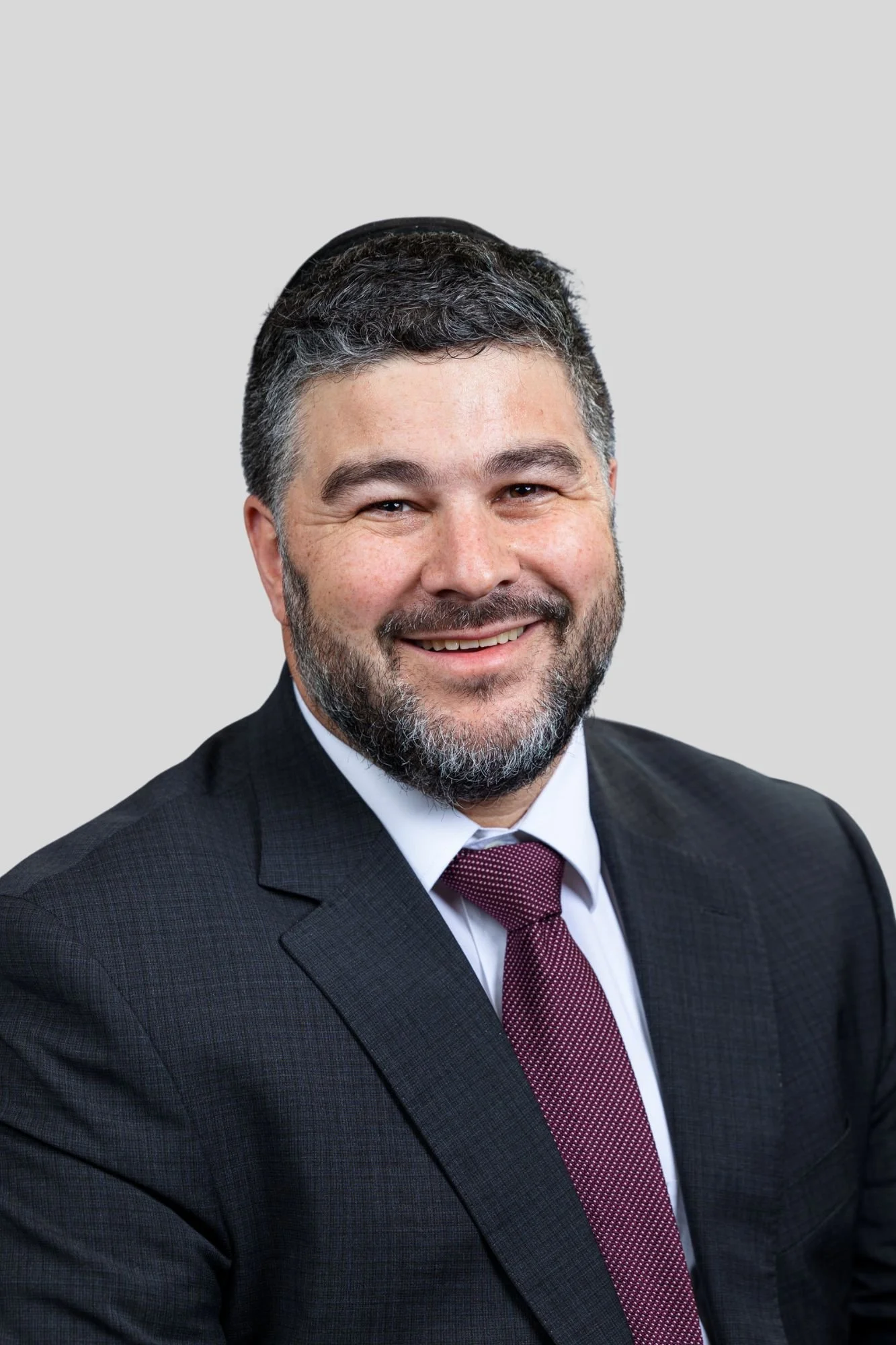 A smiling man with dark, curly hair and a beard wearing a dark gray suit, white dress shirt, and purple tie against a light gray background.