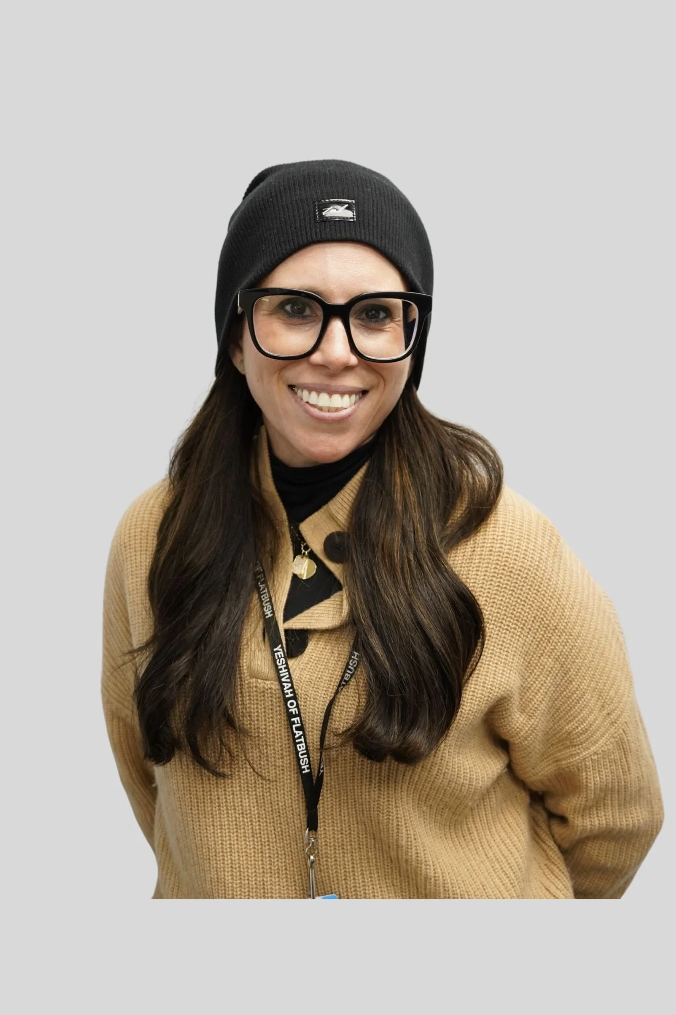 A woman with long brown hair smiling, wearing black glasses, a black beanie, a tan coat, and a black lanyard with white text, standing against a plain light gray background.