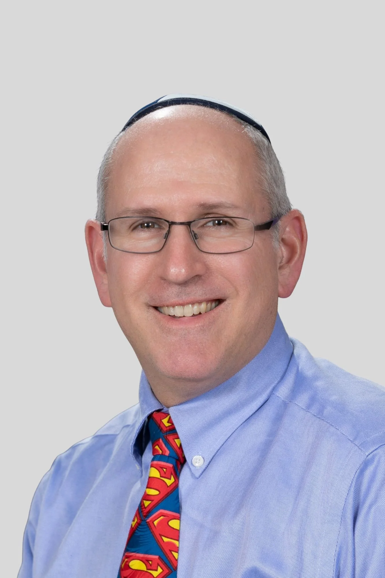 A smiling man with glasses, wearing a blue shirt, a colorful Superman-themed tie, and a yarmulke, against a plain gray background.