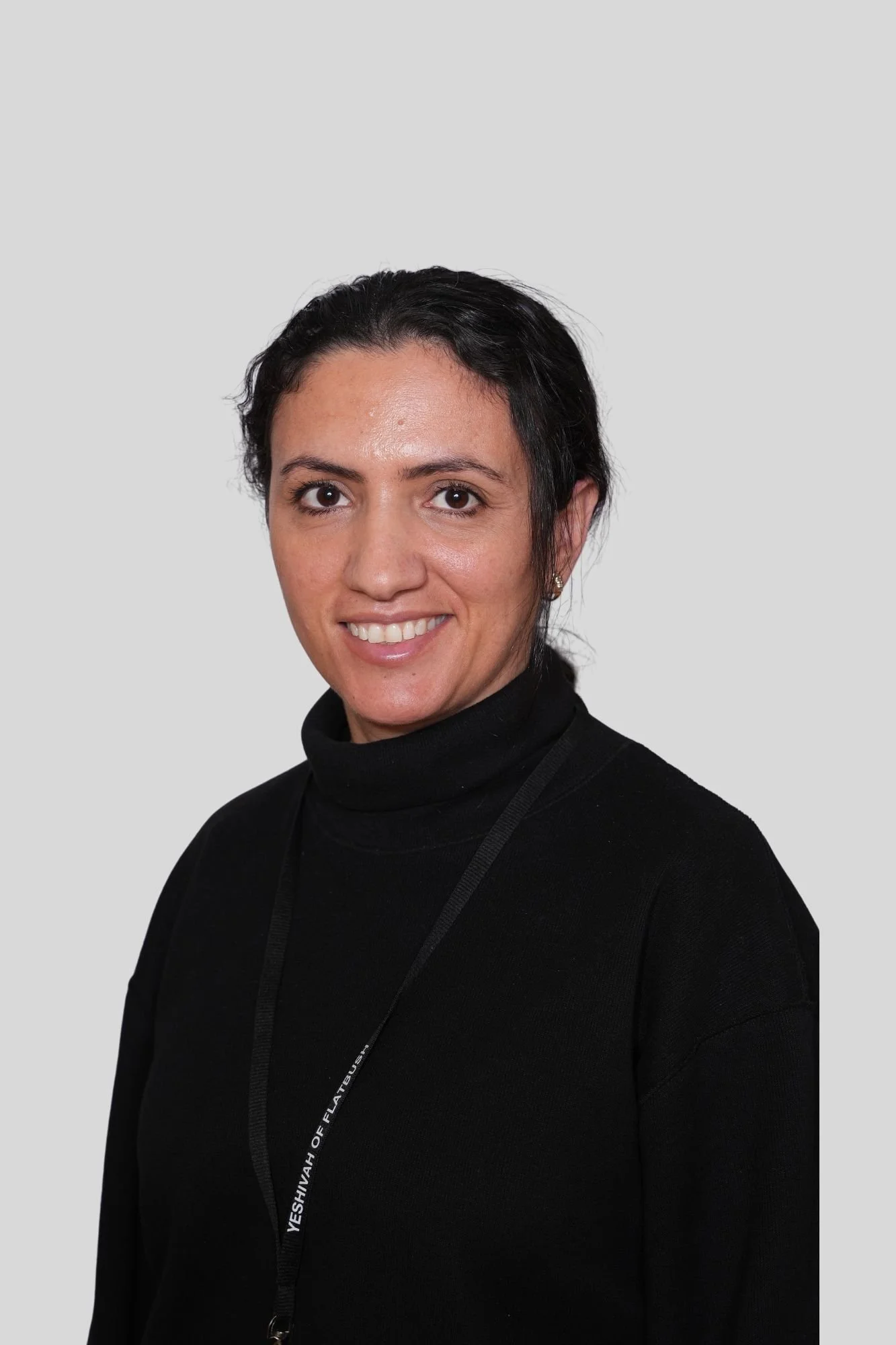 A woman with dark hair, wearing a black turtleneck and a lanyard around her neck, smiling at the camera against a light grey background.