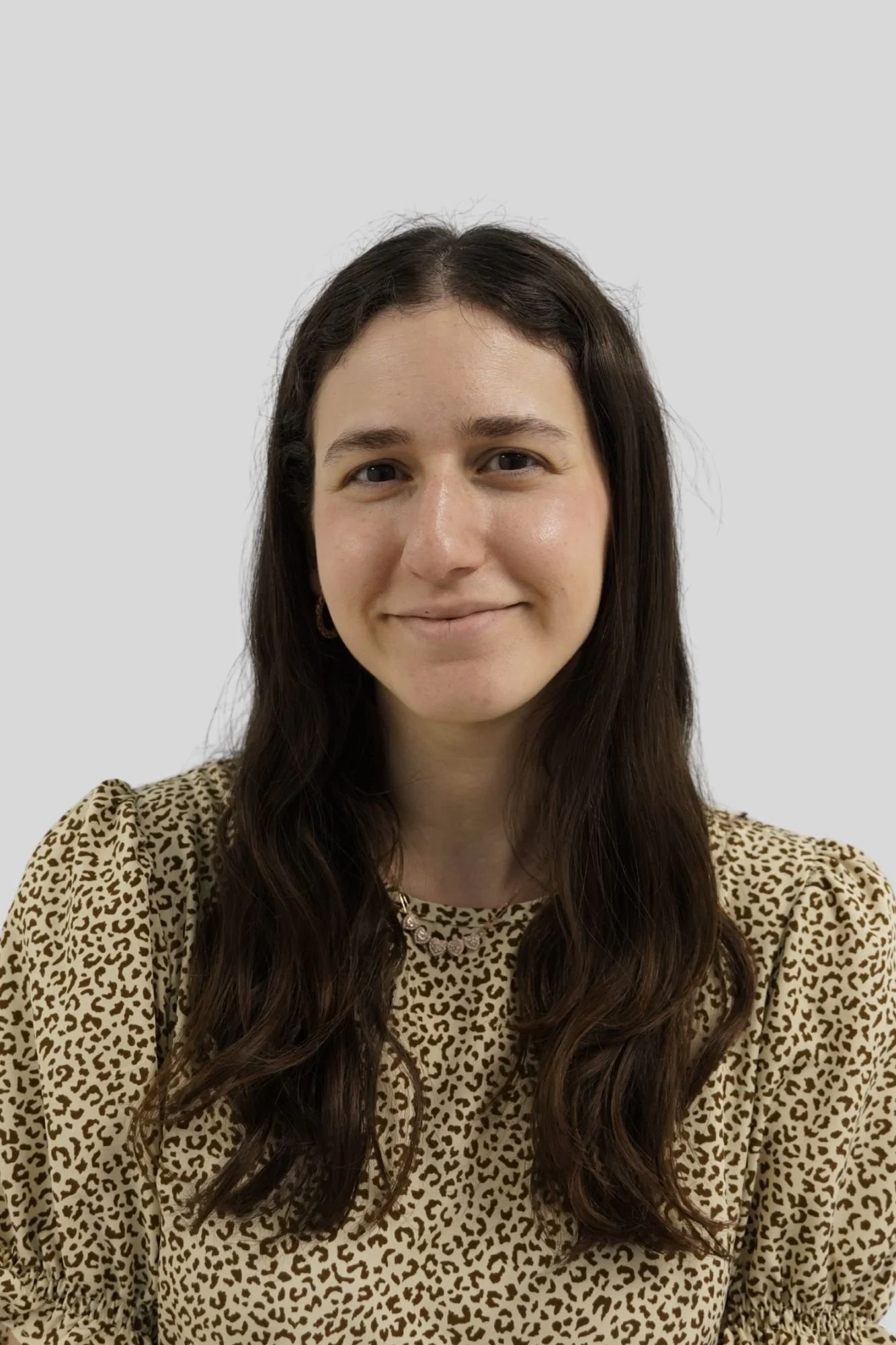 A woman with long brown hair, wearing a leopard print shirt, smiling in front of a plain light gray background.